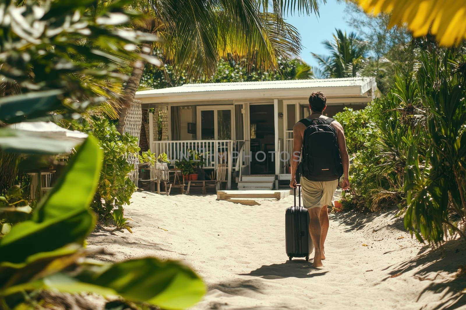 A traveler carries a black suitcase while walking on a sandy path toward a tropical beach house. Surrounded by lush greenery, this serene scene evokes feelings of relaxation and adventure in a warm, sunlit destination.