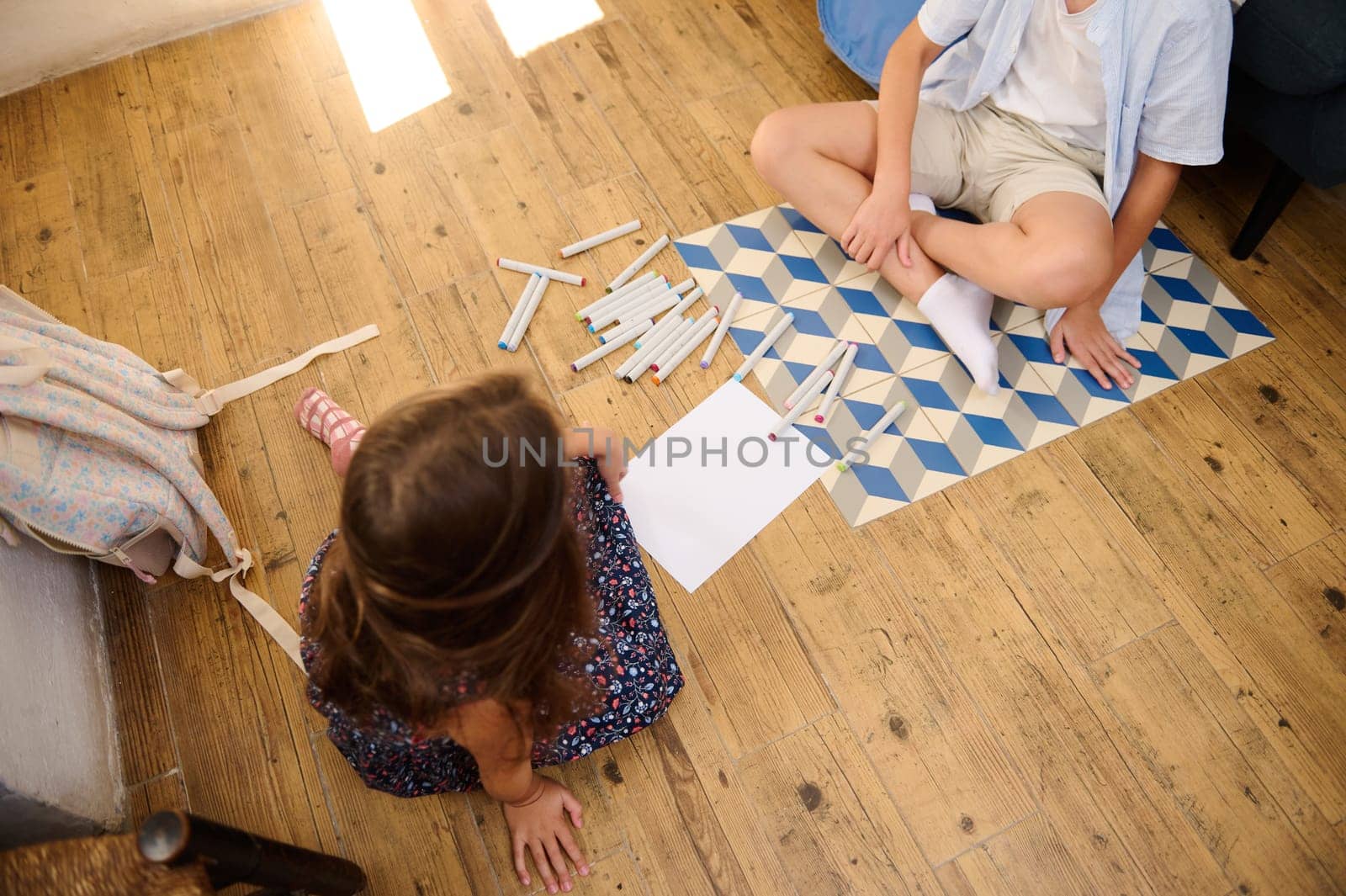 Two children enjoying creative activities, drawing with markers on a wooden floor. The scene captures teamwork and artistic expression.