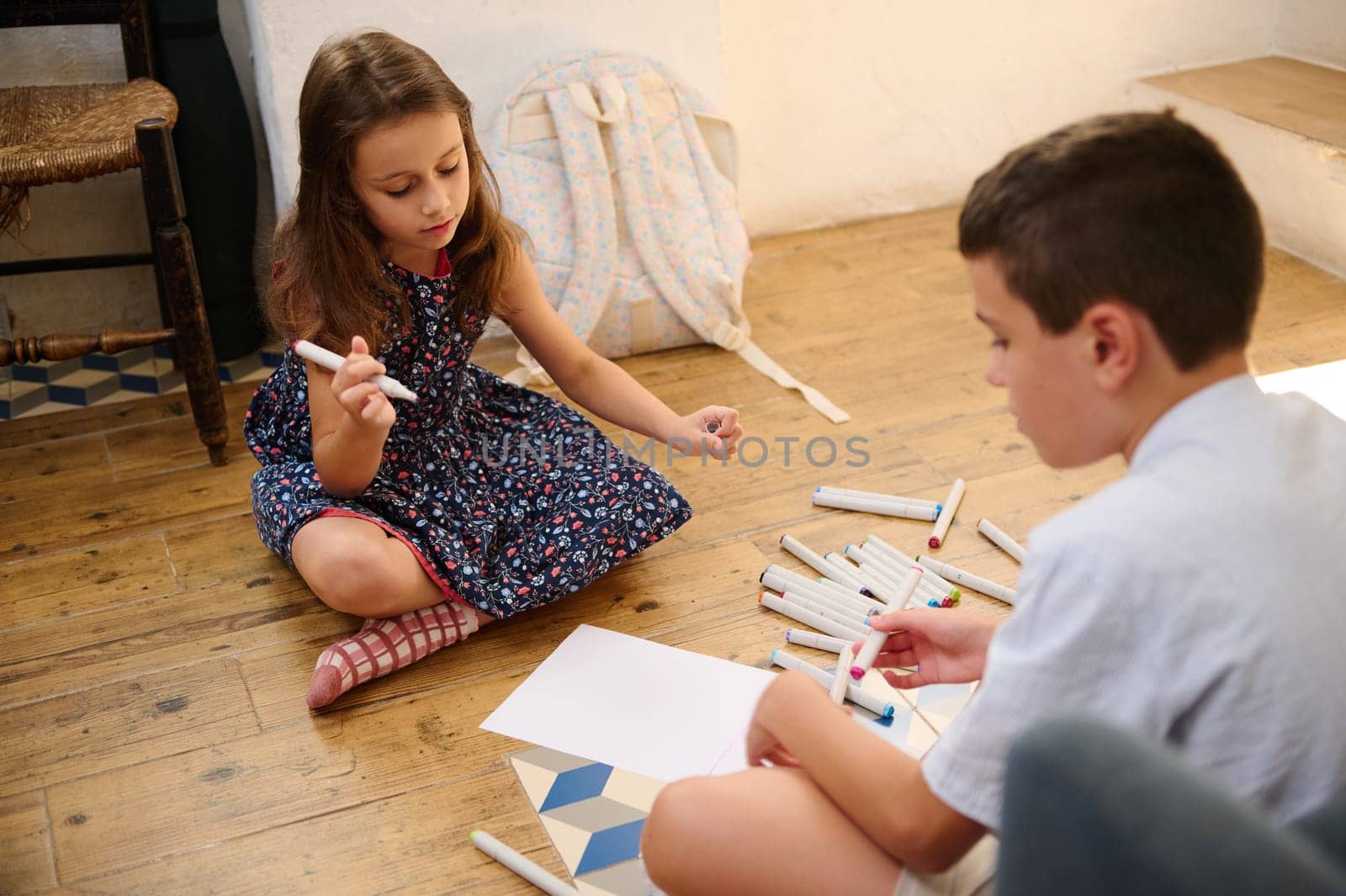 Two children are seated on the wooden floor, engaging in creative drawing activities using markers, showcasing their colors and imagination.
