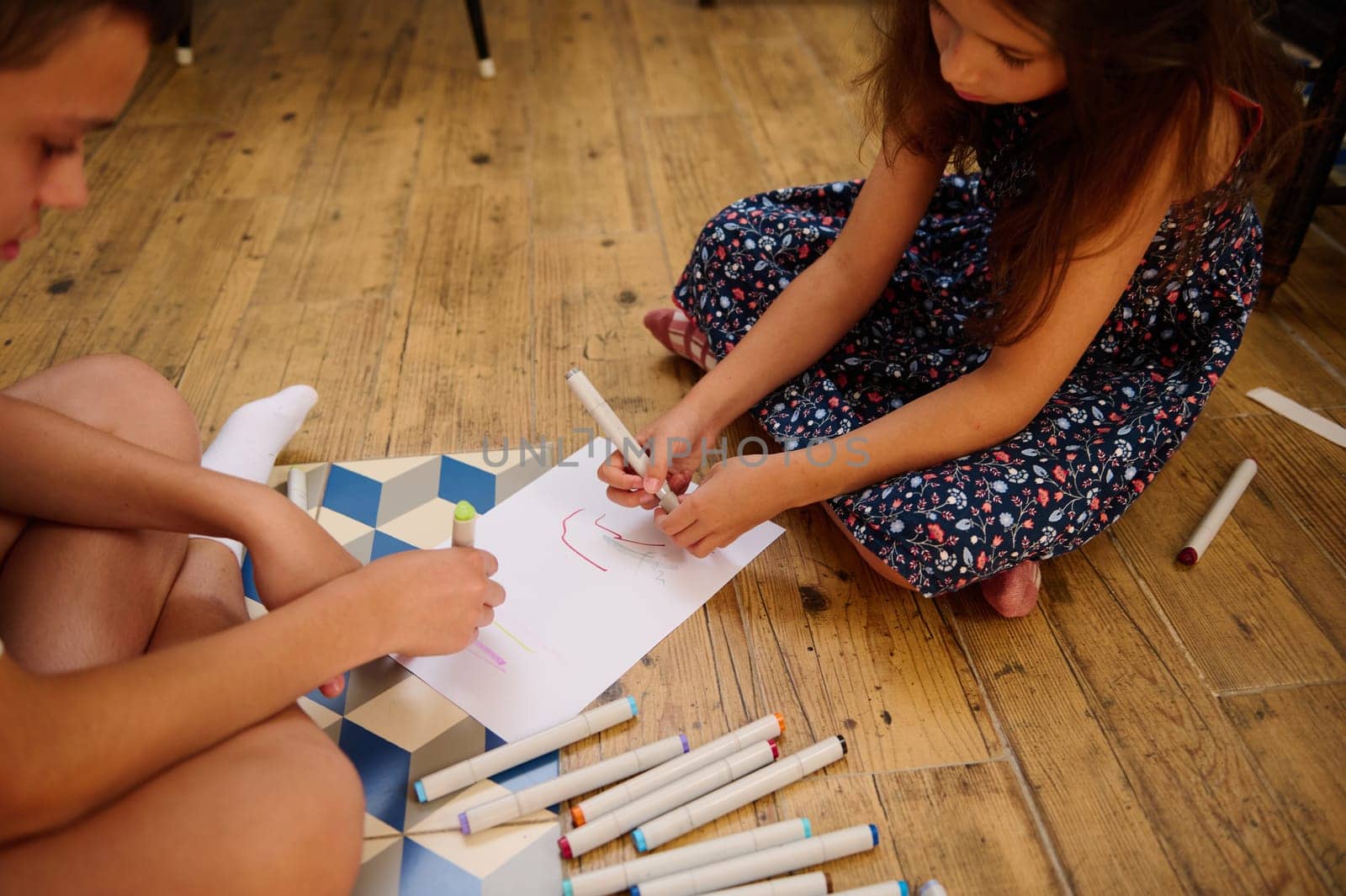 Children sitting on a wooden floor using colorful markers to draw on white paper, fostering creativity and collaboration.