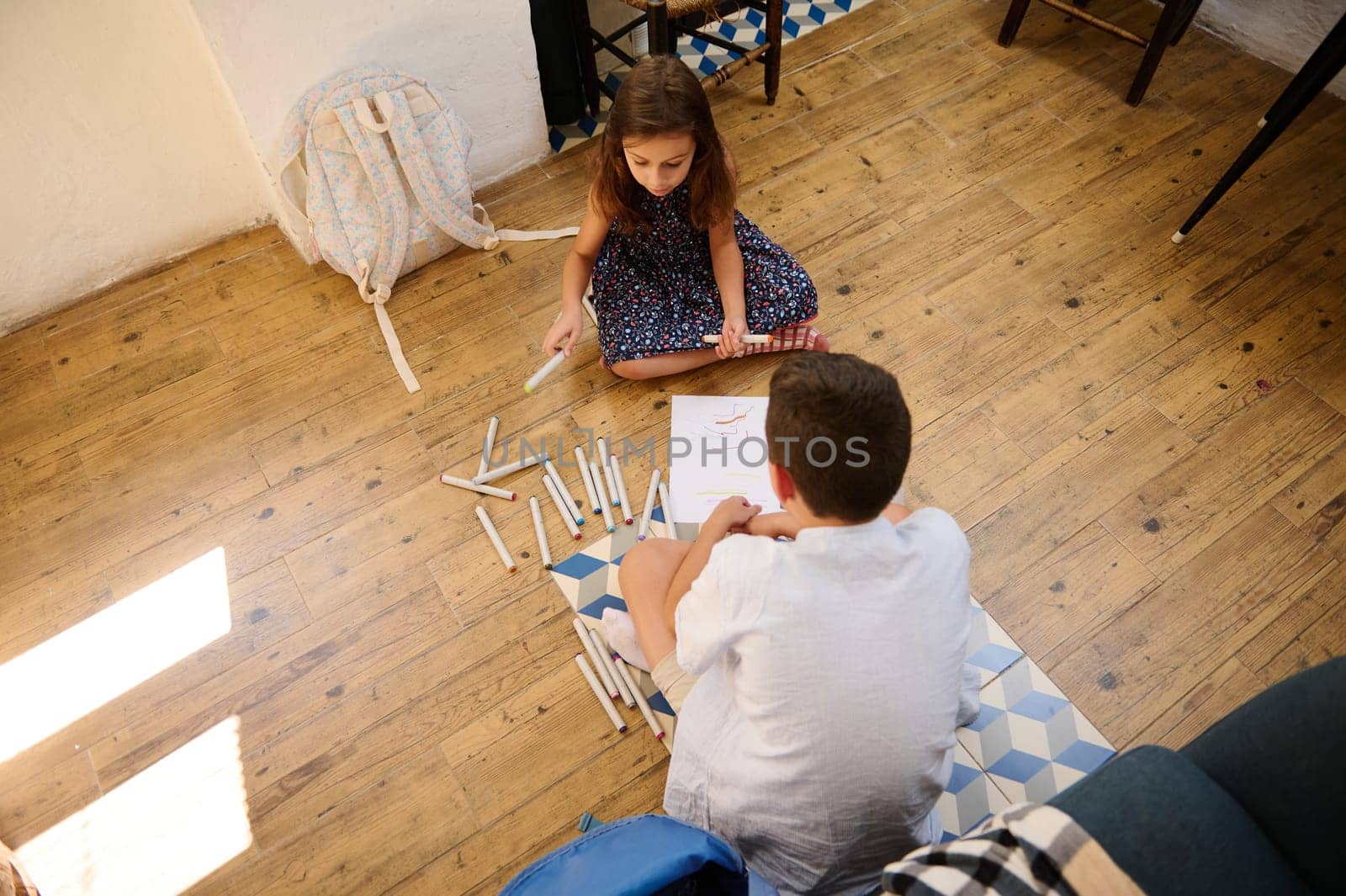 Two children sitting on a wooden floor, drawing with colorful markers, fostering creativity and cooperation.