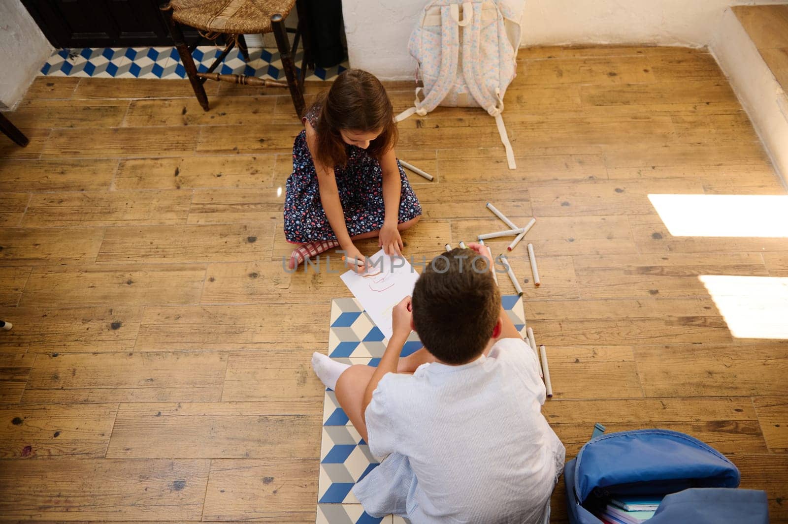 Two children collaborating on a craft project seated on a wooden floor. Their materials, including colored pencils, are scattered, showcasing a moment of creative and educational bonding.