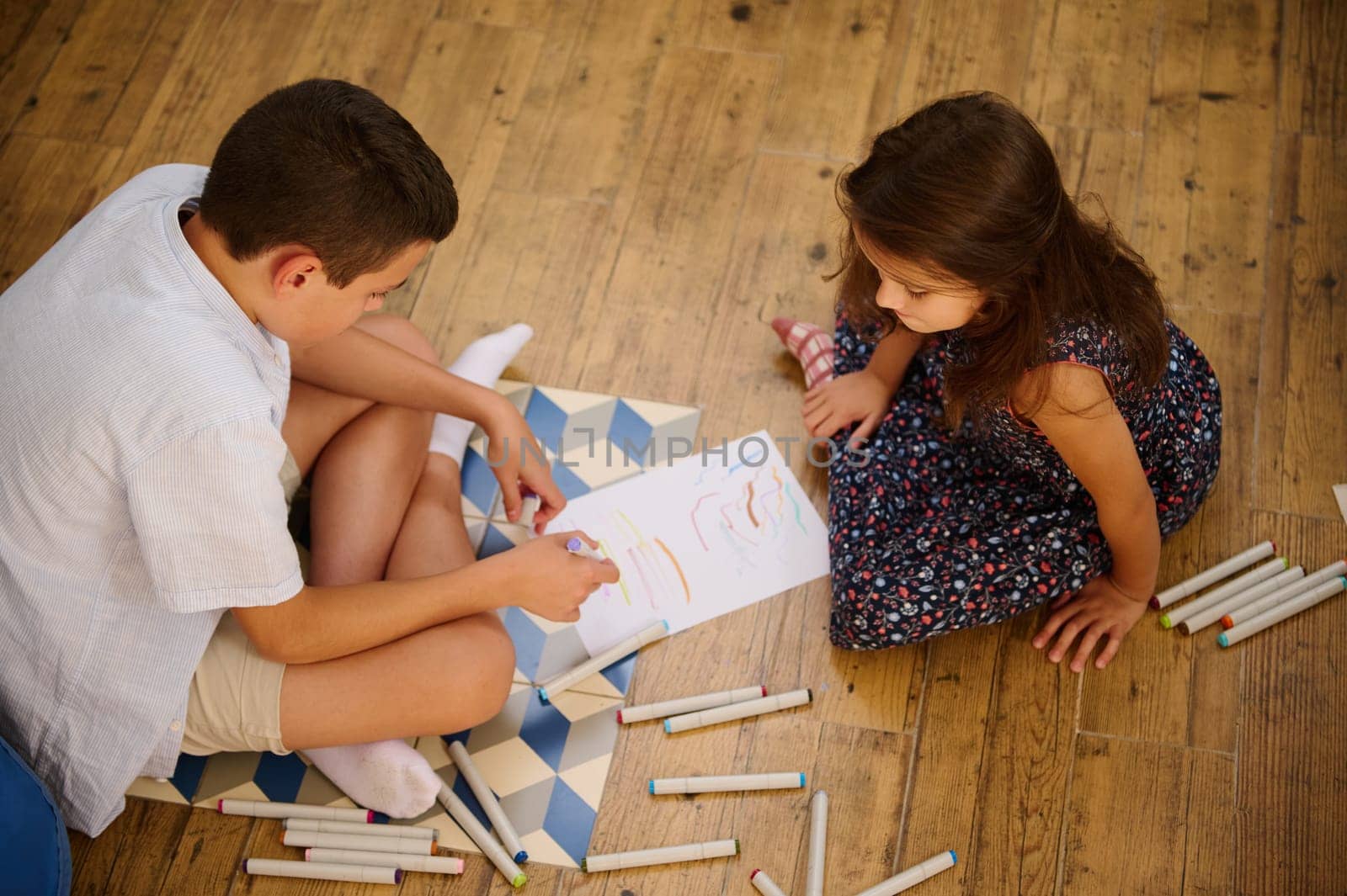 A boy and girl create art on paper while sitting on a wooden floor, using colored markers, sharing creativity and bonding.