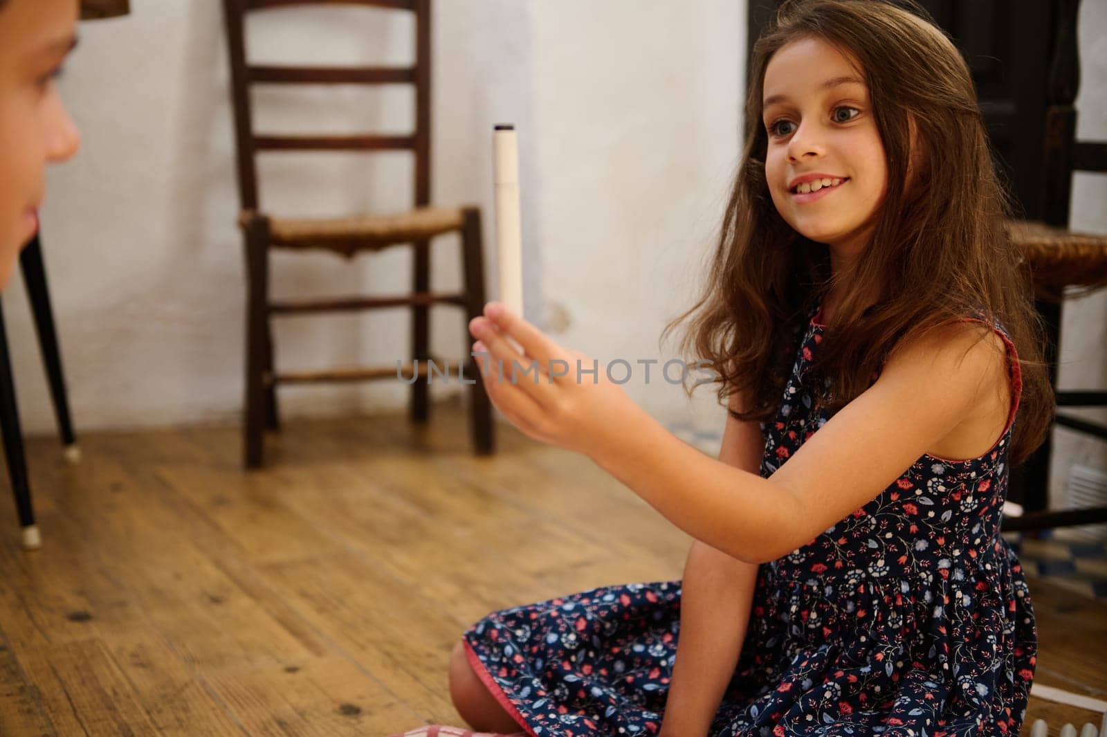 A cheerful girl in a flowered dress smiling and holding a pen during indoor casual play.