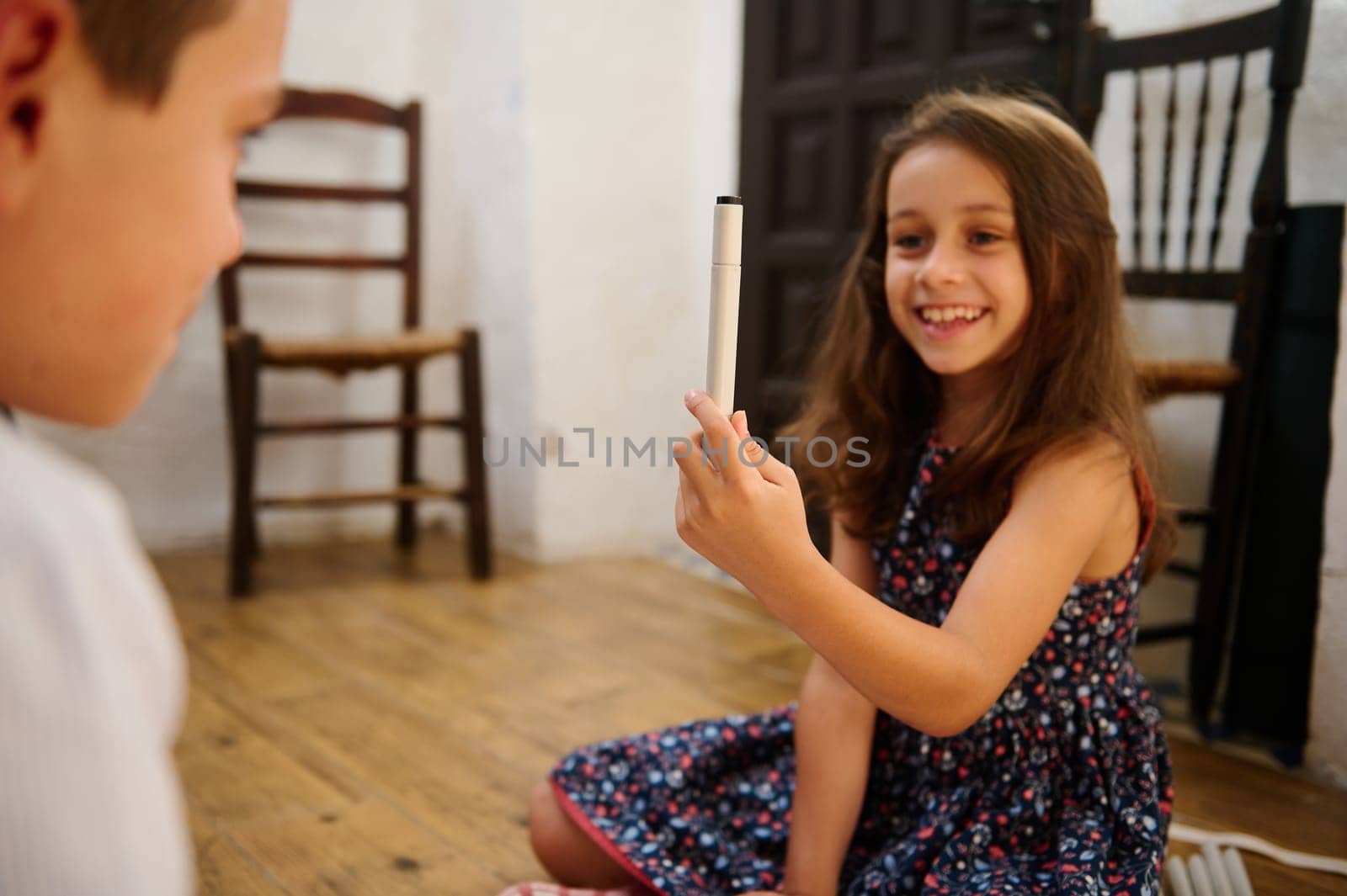 A young girl in a floral dress smiling while holding up a pen indoors, sharing joy.