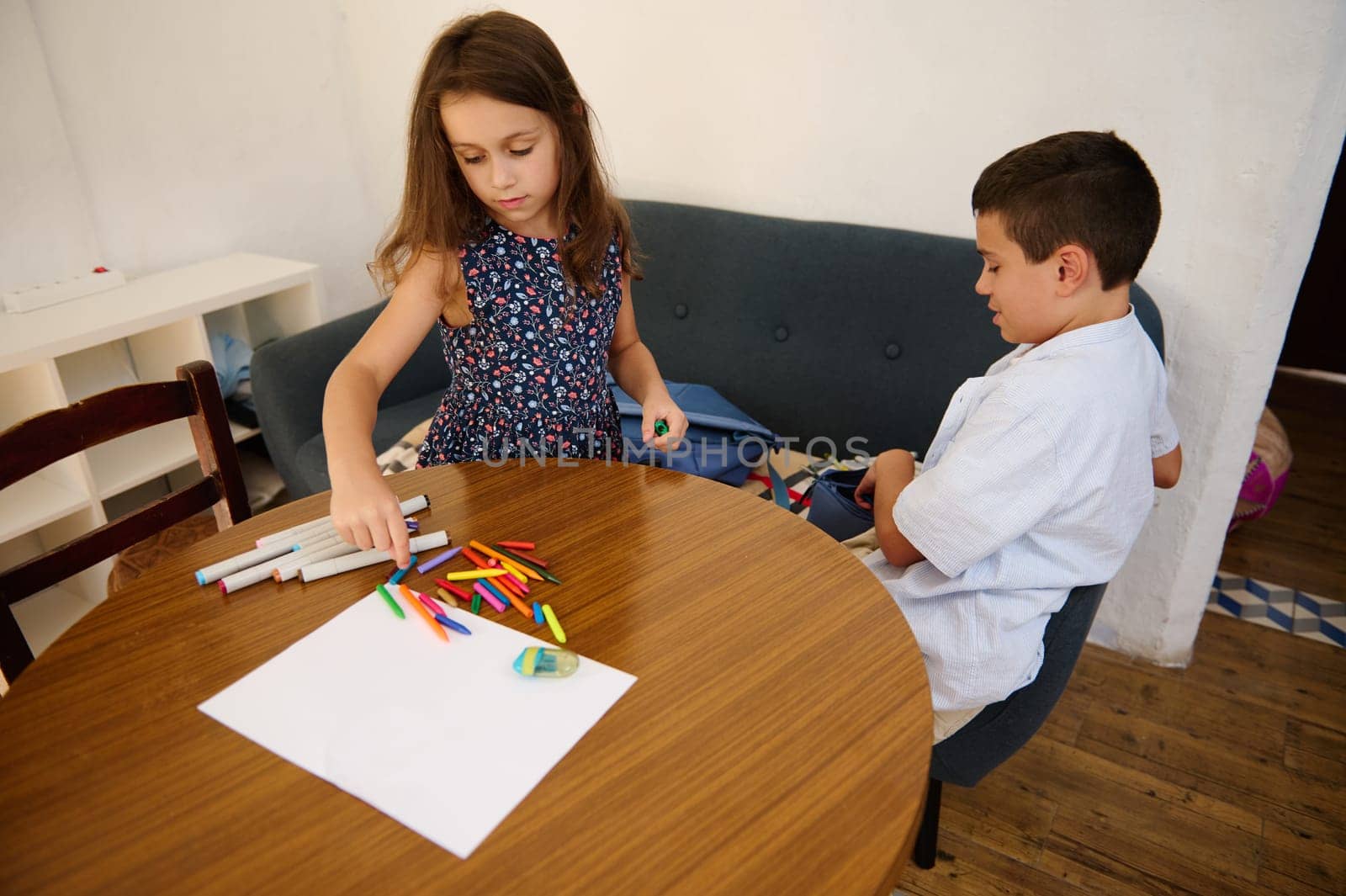 Two children sit at a wooden table working on creative crafts with markers, pencils, and paper. The scene emphasizes creativity, childhood learning, and the joy of art-making activities.