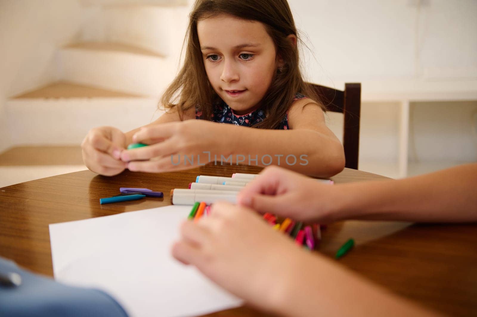 Young girl sitting at a table drawing with colored markers, expressing creativity and focused on her activity.