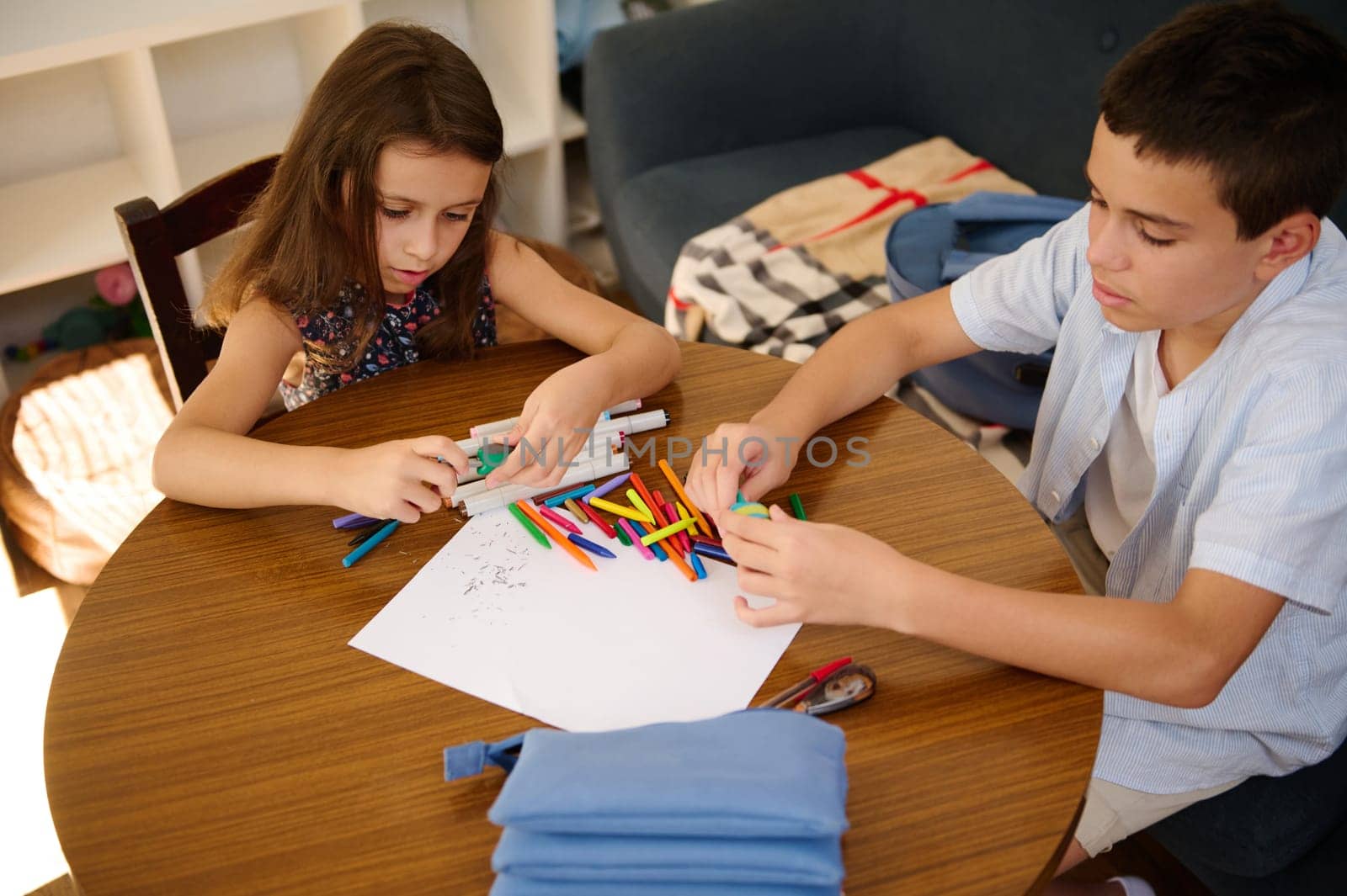 Two children sit at a table engrossed in a collaborative creative activity, sorting colorful markers and making art on blank paper.