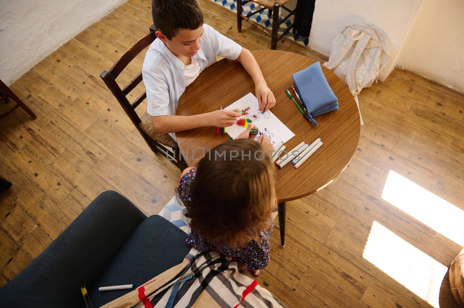 Two kids engage in a creative activity, coloring and drawing, at a wooden table in a cozy indoor setting. A mix of art supplies and bright sunlight enhance the vibrant atmosphere.