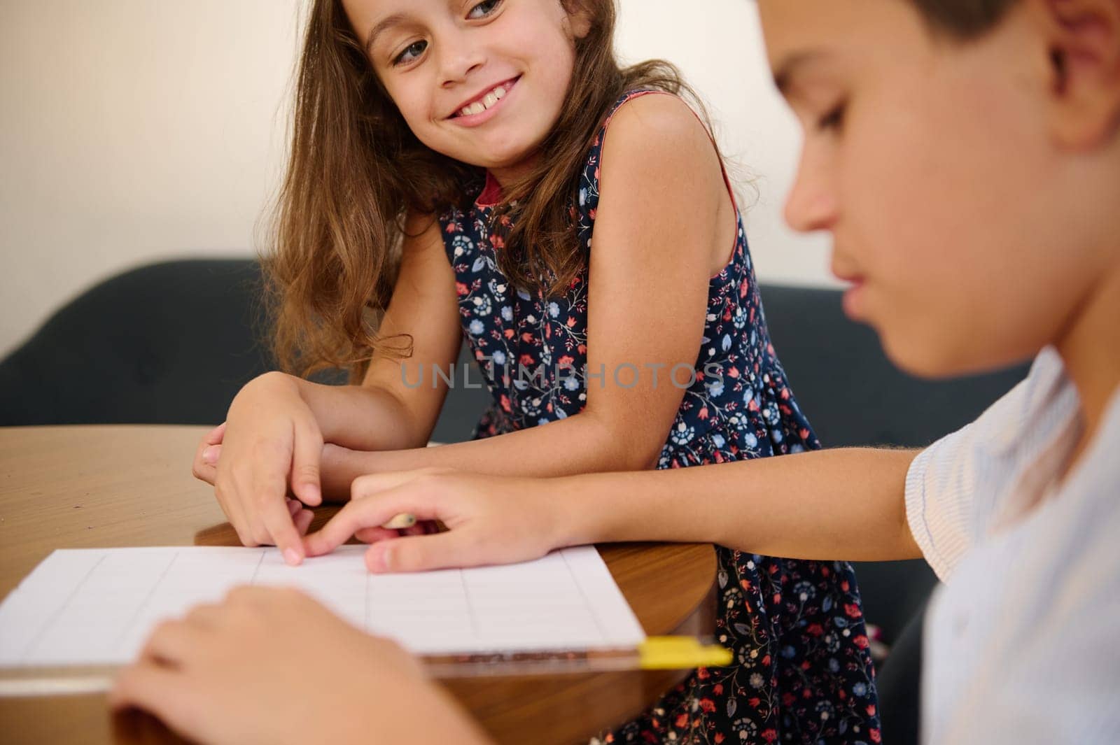 Two young children sitting at a table, collaborating on a school project with interest and focus. The girl smiles warmly as the boy works attentively.