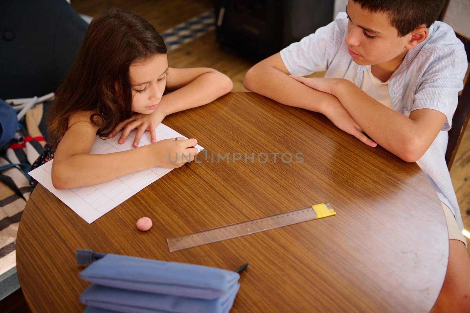 Two children collaborating at a wooden table, one writing on a paper while the other observes, emphasizing education, learning, and teamwork in a warm indoor environment.