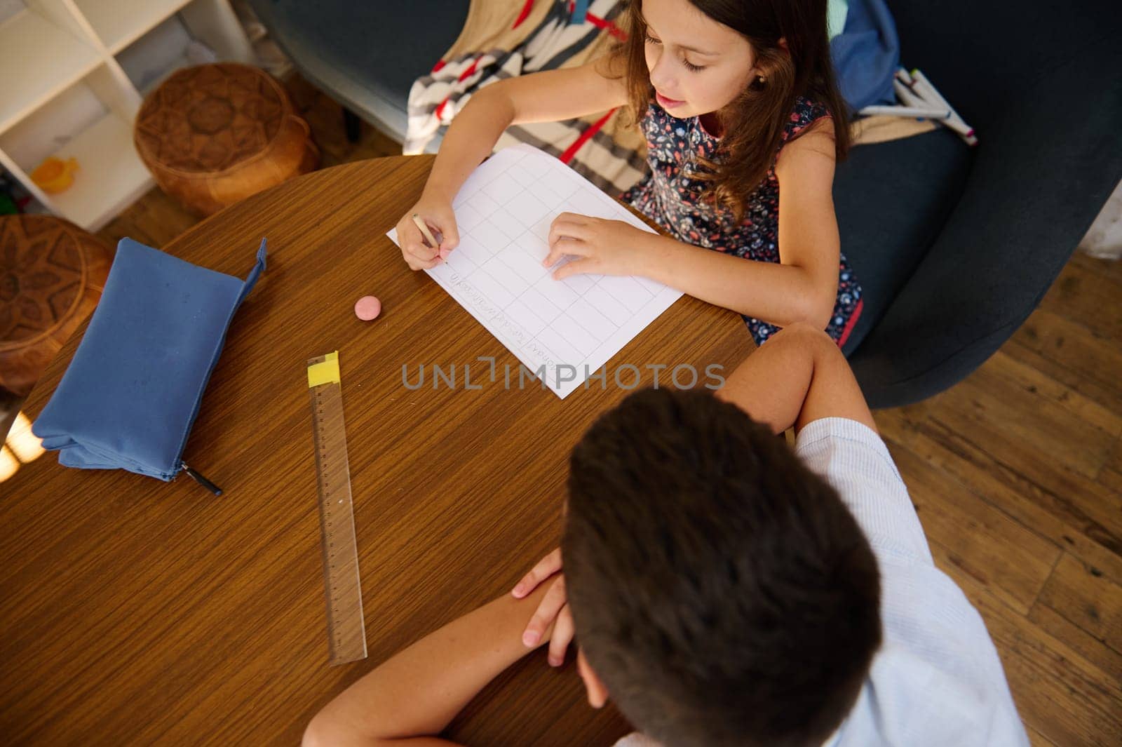 A young girl and boy working on homework together at a wooden table in a colorful classroom setting.