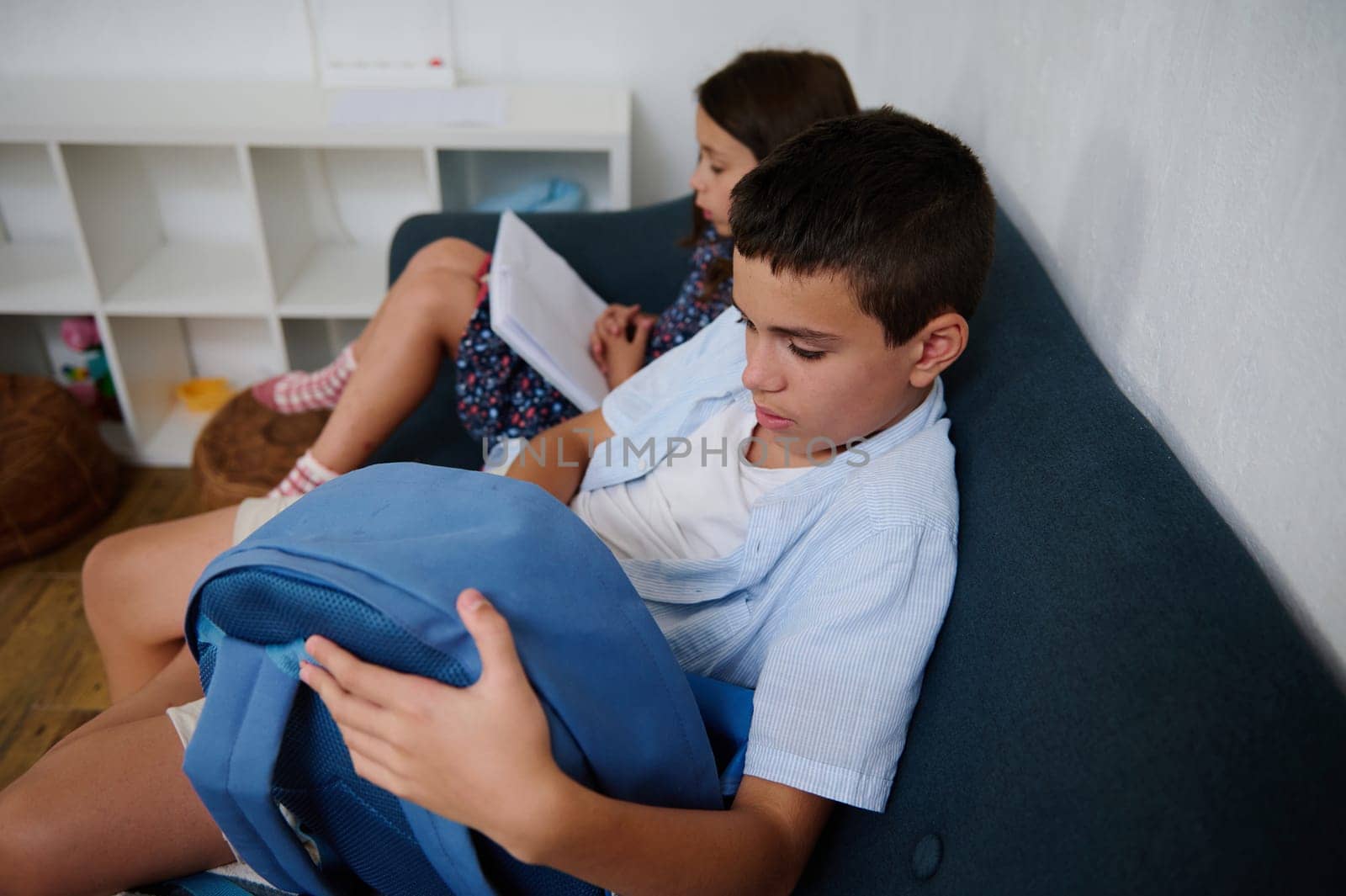 Two children sitting on a couch, one reading a book while the other looks into a bag. A cozy and calm indoor scene highlighting study and leisure.