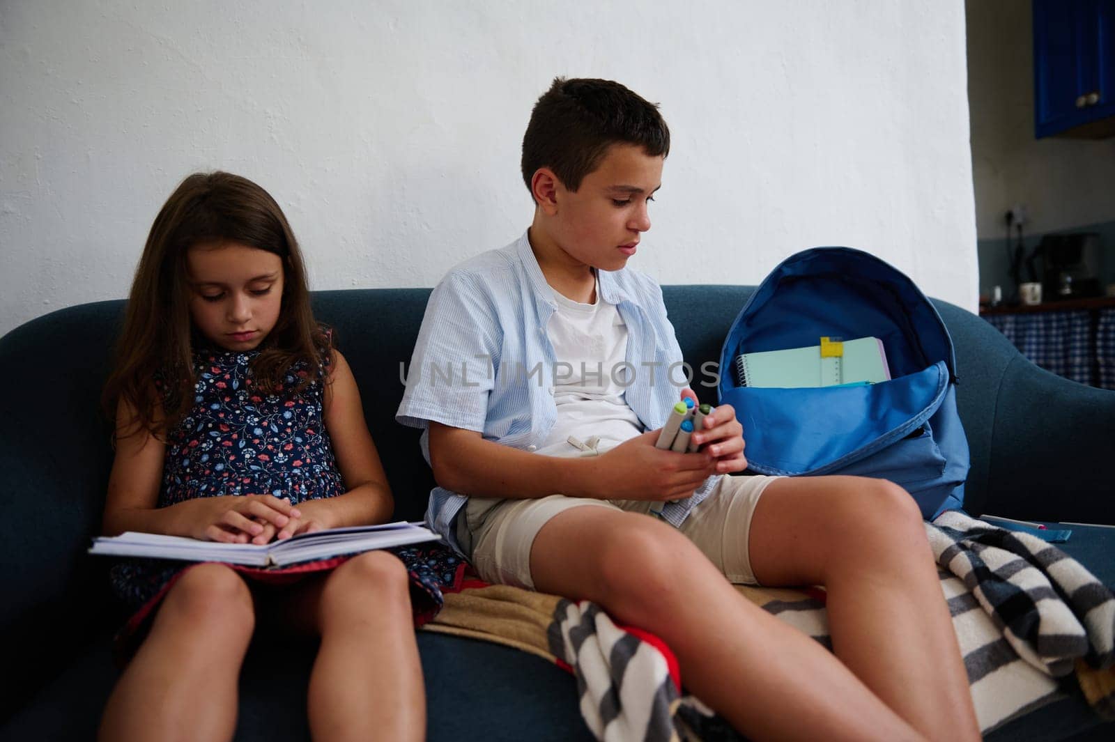 A boy and girl sit on a sofa reading books and organizing supplies with focus and attention.