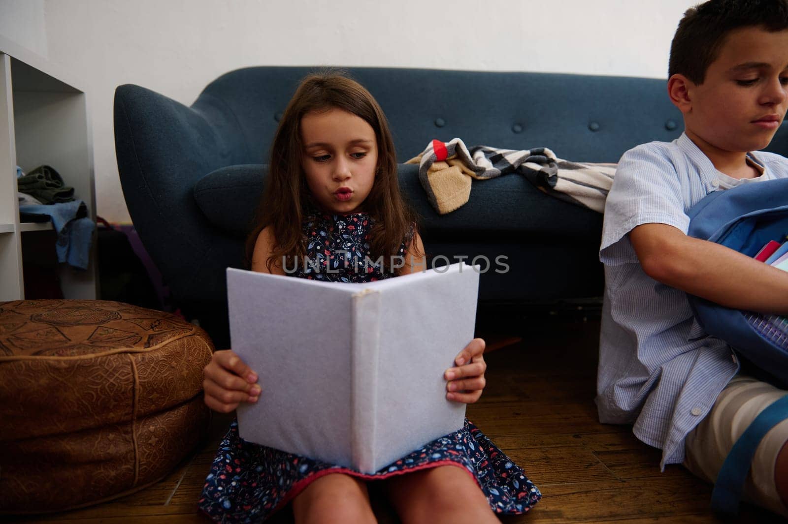 A young girl sitting on the floor reads a book while her brother organizes his things in a home setting. The scene exudes focus and calm.