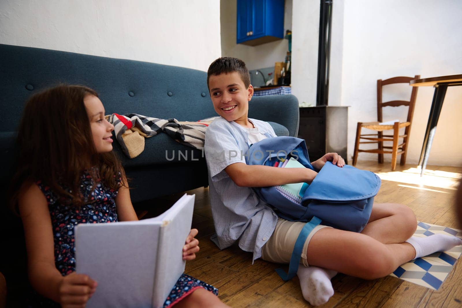 A brother and sister studying and interacting while sitting in a cozy home environment with books and backpacks.
