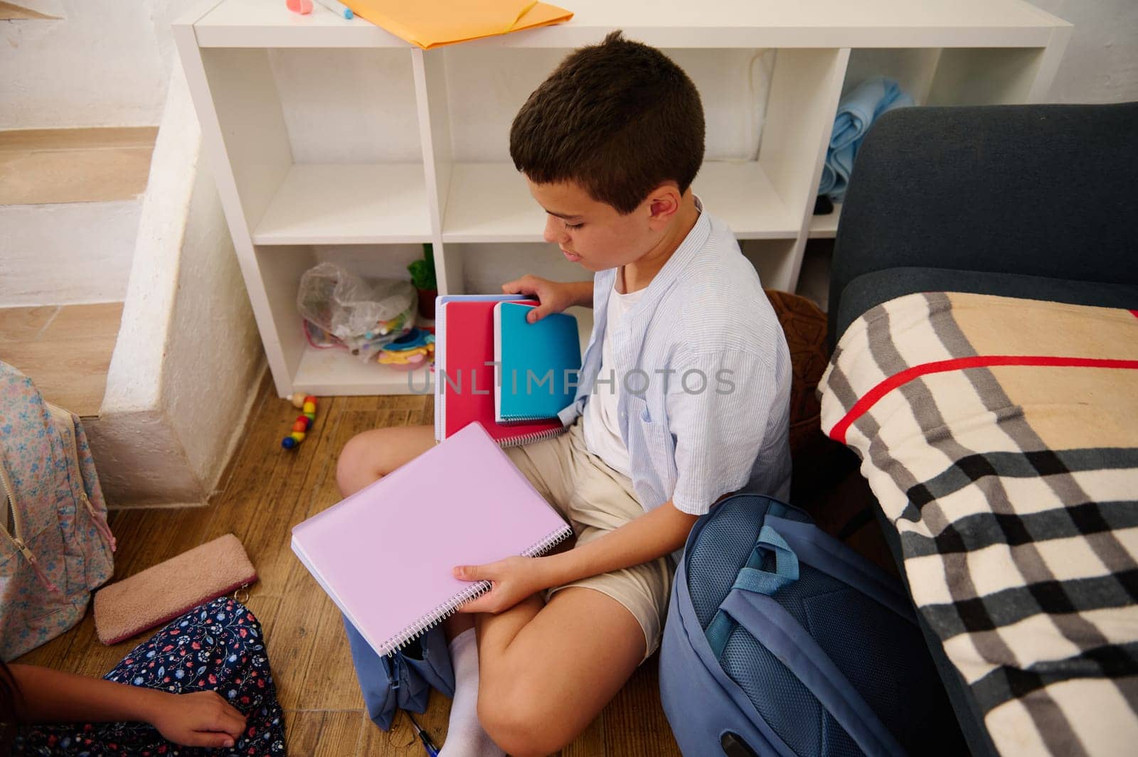 A young boy sits on the floor, sorting notebooks from his backpack into neat stacks, conveying preparation and focus in a home setting with shelves and toys in the background.