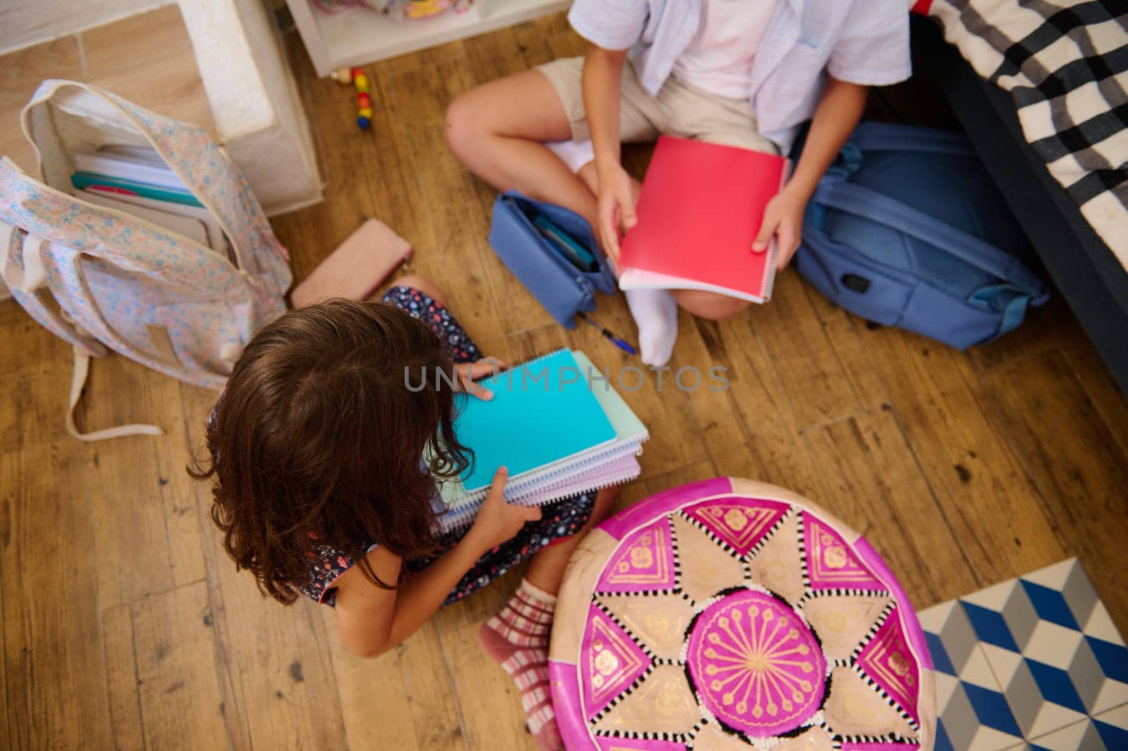 Two children sit on a wooden floor, holding notebooks and sharing materials, fostering collaboration and creativity.