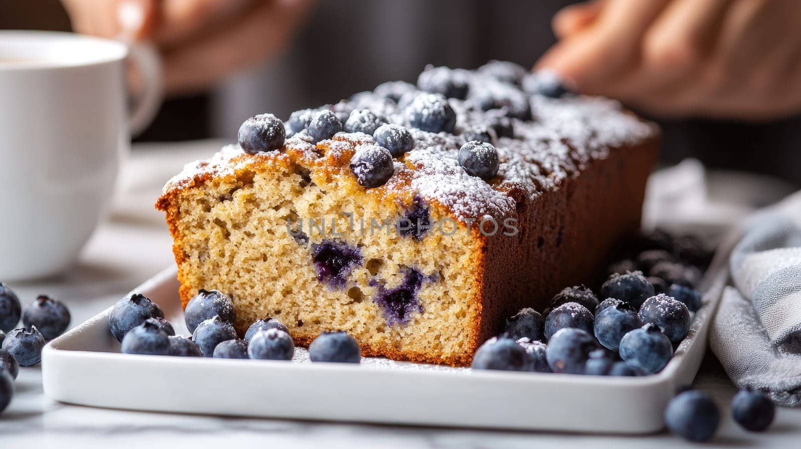 Freshly baked blueberry loaf cake is garnished with powdered sugar and surrounded by blueberries on a white plate, perfect for a comforting morning treat with coffee.