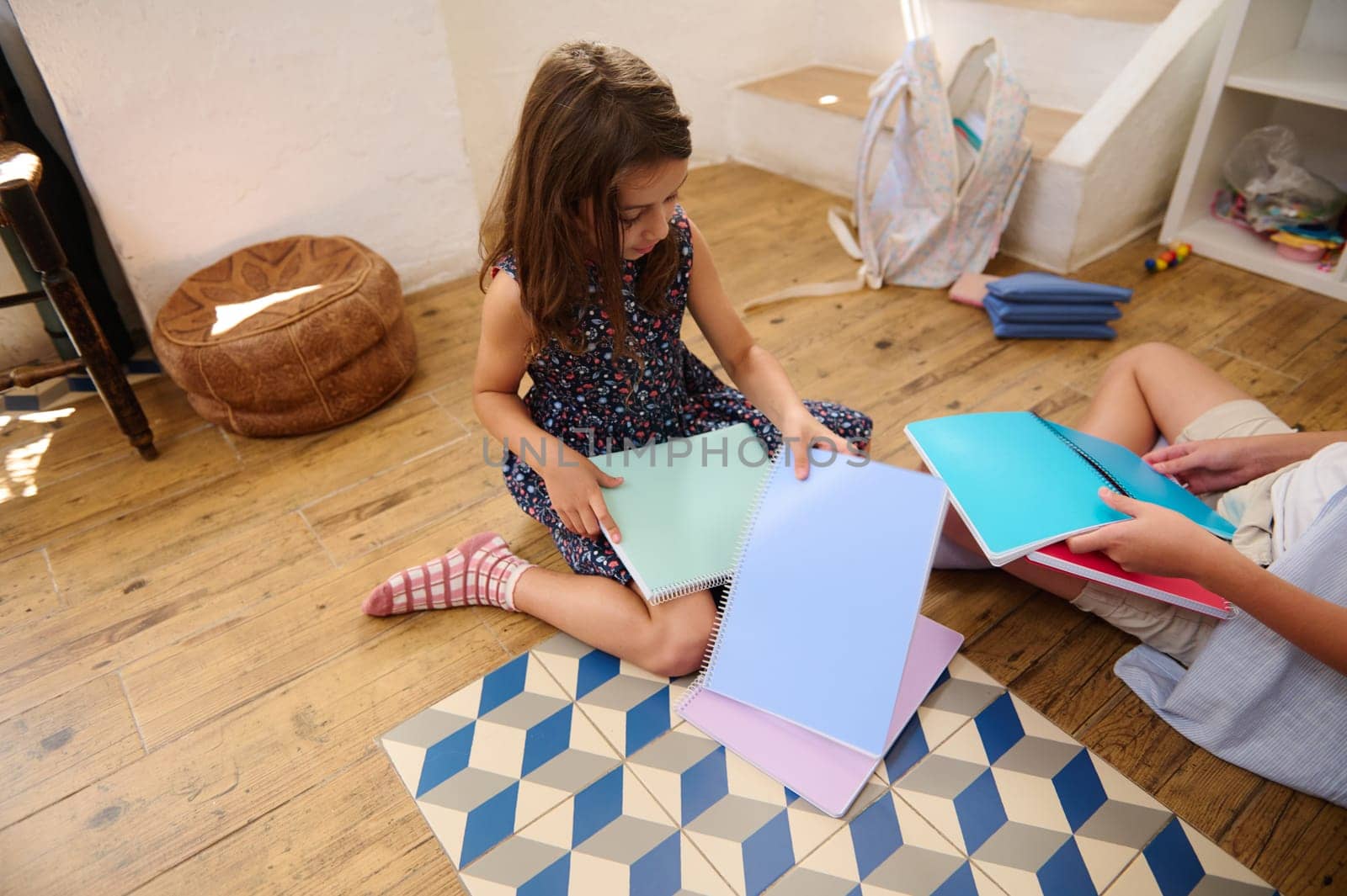 Two children engaged in a study session with colorful notebooks, sitting on a wooden floor in a cozy room. A casual educational and creative environment.