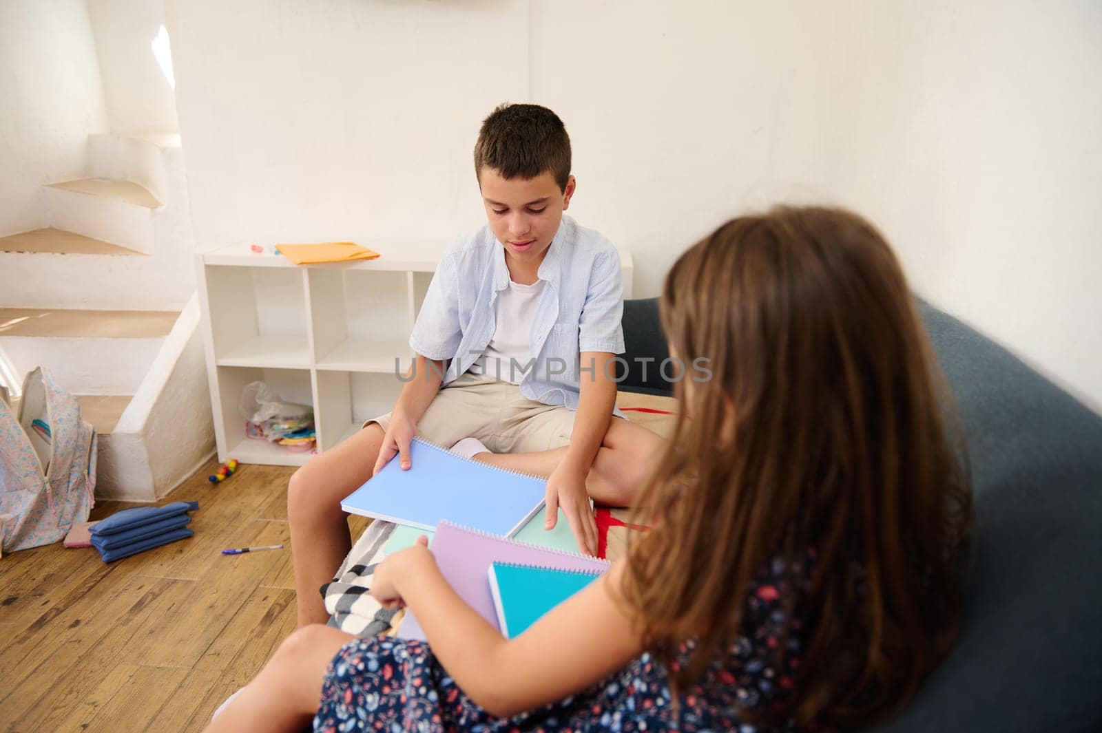 Two children collaborate on a sofa, sharing notebooks in a cozy environment. Captures themes of learning, education, teamwork, and childhood in a relaxed and informal setting.