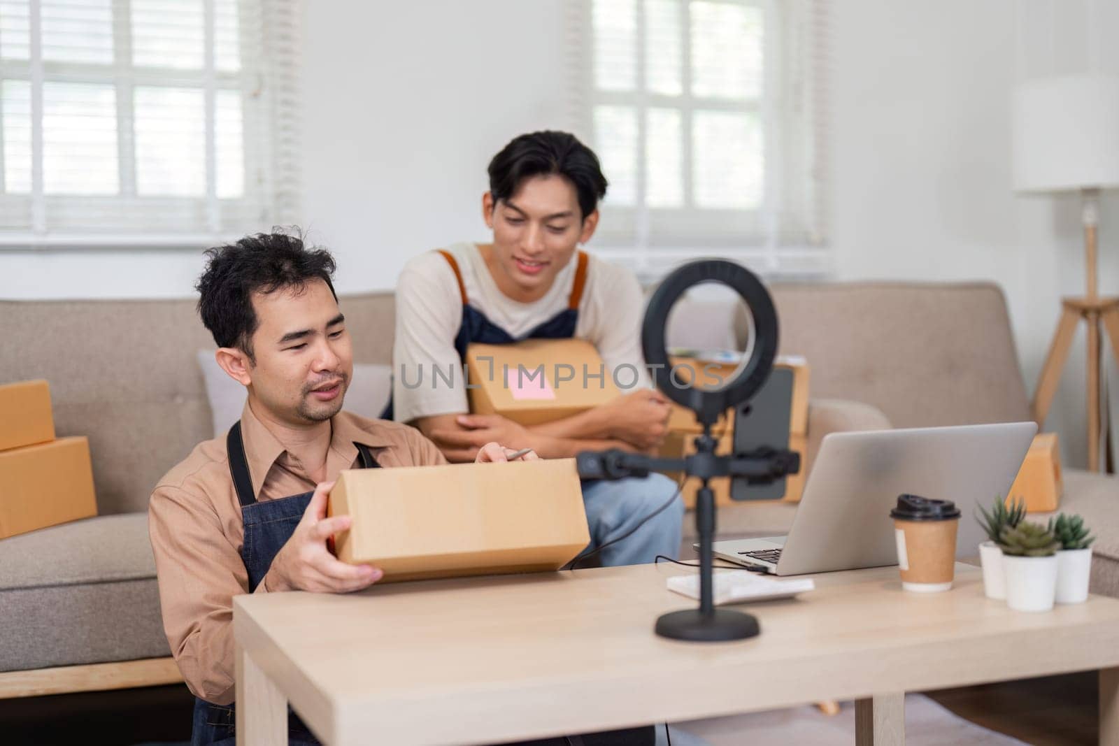 Two young Asian entrepreneurs getting ready for an unboxing session in their home office, holding packages and setting up equipment to engage with their audience.