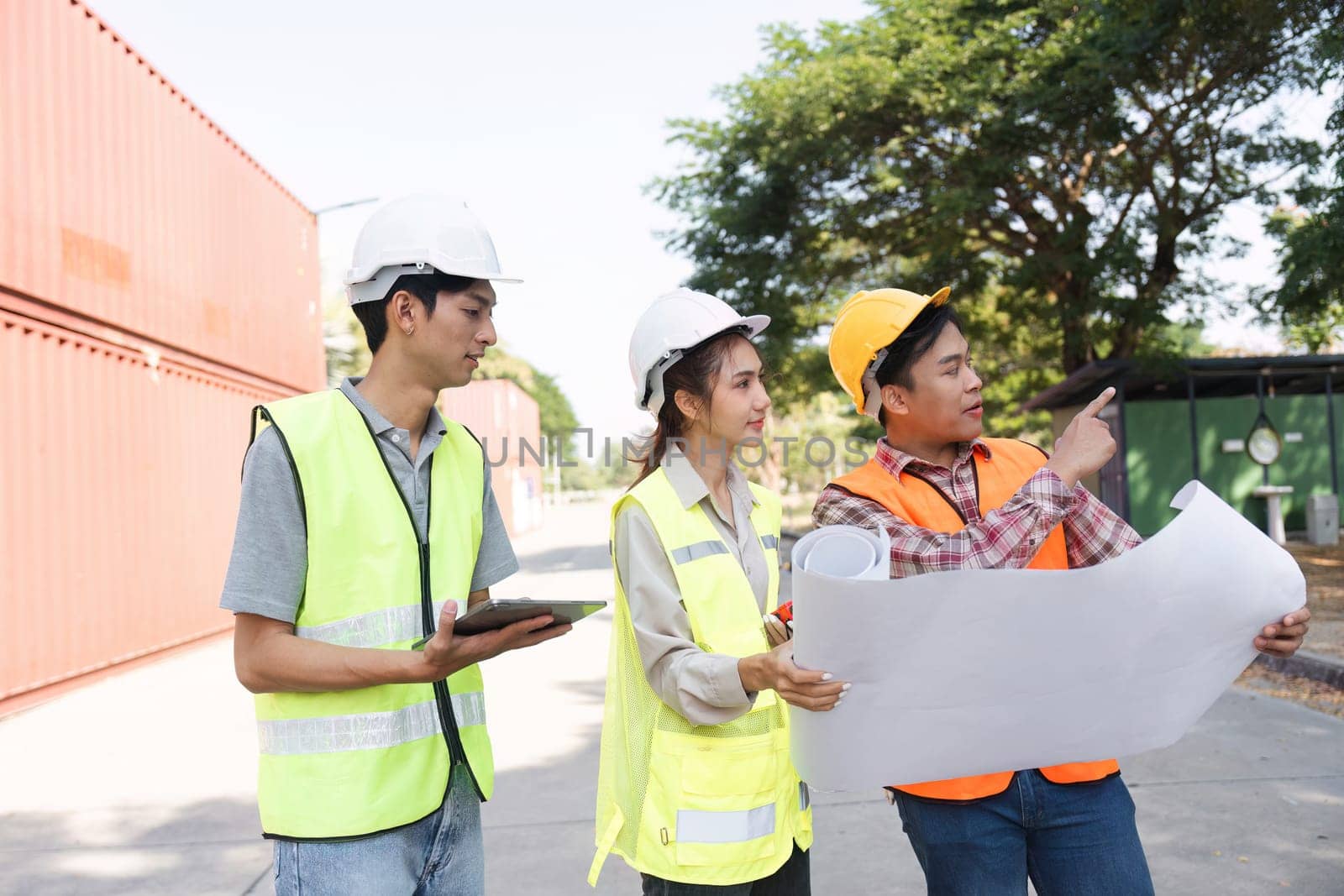 Three construction professionals, including an engineer, a project manager, and a worker, analyzing blueprints and discussing plans at an active construction site.