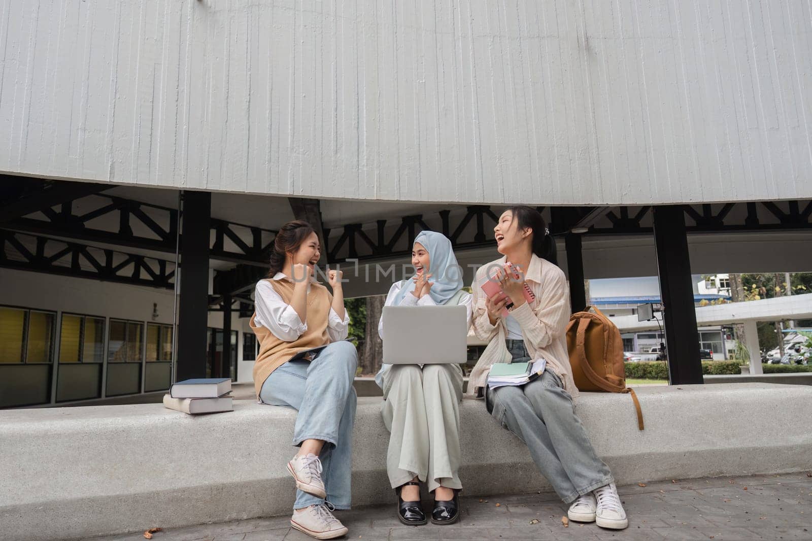 Diverse female students engaged in a lively conversation, smiling and sharing ideas while using a laptop and surrounded by books in a university campus setting.