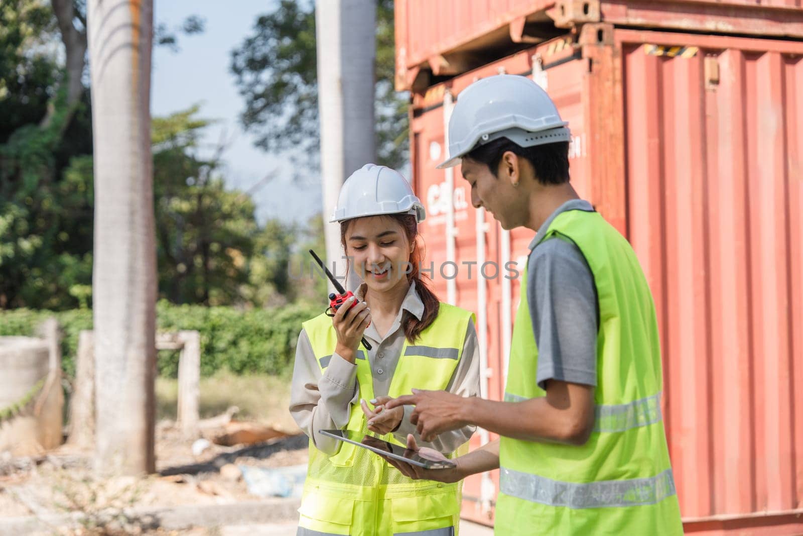 Two construction engineers use radios to communicate project updates while reviewing plans near shipping containers, emphasizing teamwork and coordination.