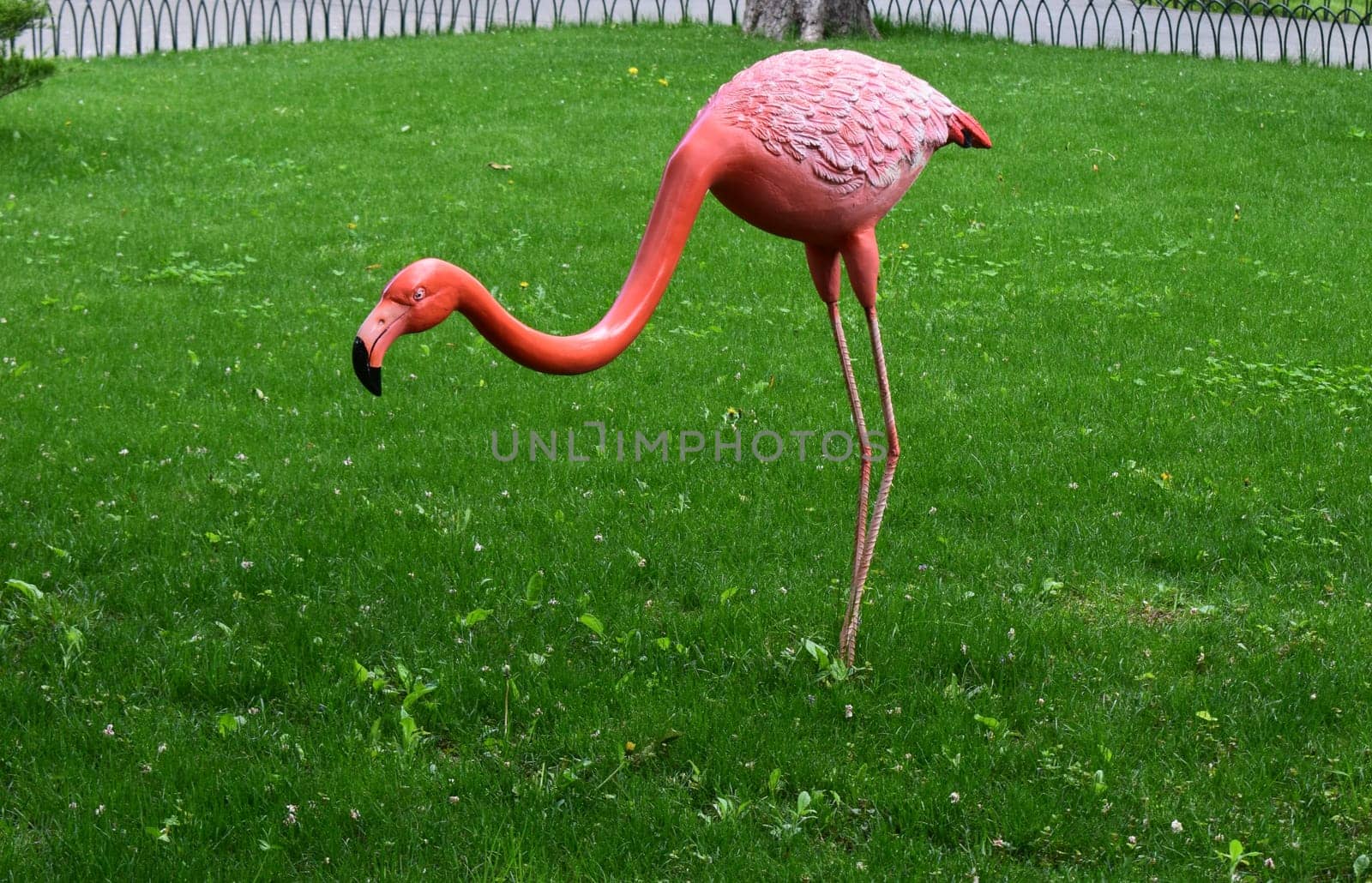 Kharkov, Ukraine, 14 May 2024. A figure of a pink flamingo on a green lawn. The figure has a long curved neck, a detailed pink body with feather texture, and slender legs. In the background, there is a fence and some bushes.