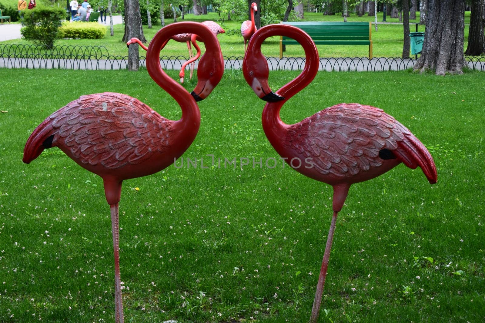 Kharkov, Ukraine, 14 May 2024. Two red plastic flamingos placed in a green park. They are facing each other, creating a heart shape with their necks. In the background, you can see more flamingos, trees, bushes, green benches, a litter garbage can and people walking along the park path.