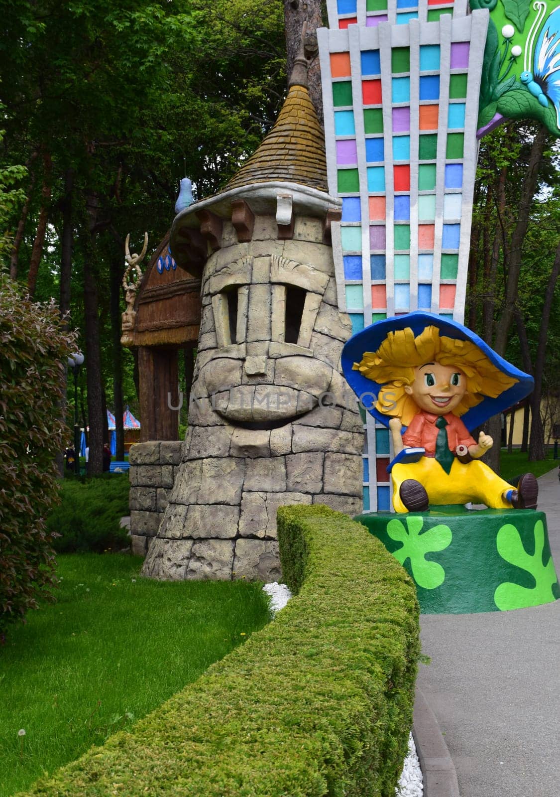 Kharkov, Ukraine, 14 May 2024. A colorful scene in an amusement park. The smiling figure in the form of a stone tower is accompanied by a sculpture of a cheerful character dressed in yellow clothes and a big blue hat (Neznayka). Behind her is a structure with a checkered pattern in bright colors. All around is greenery, manicured hedges and trees, creating a playful and whimsical atmosphere.