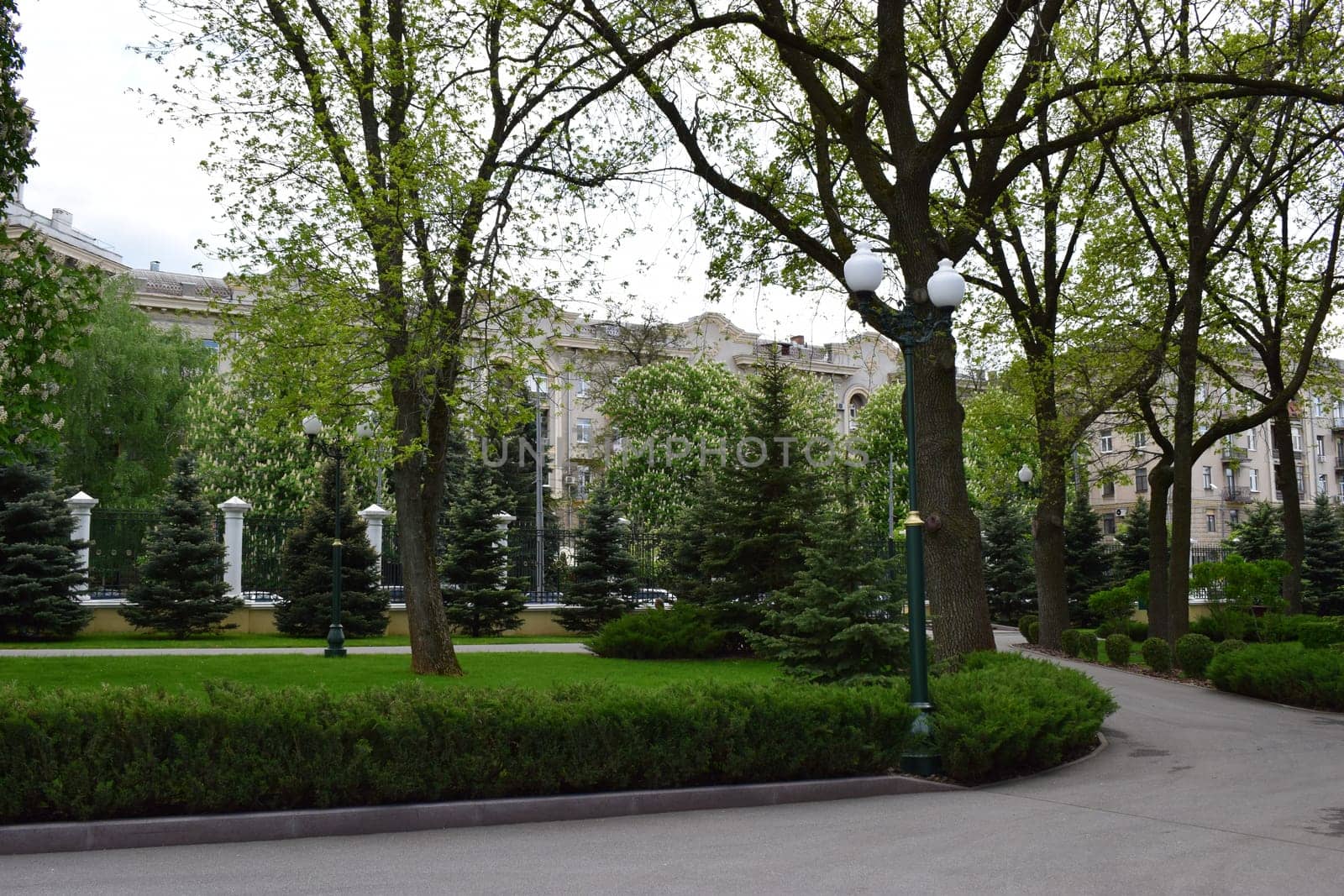 Kharkov, Ukraine, 14 May 2024. An urban park with paved walkways lined with bushes and trees. There are several elegant lampposts and large residential buildings visible through the trees. The setting is green and well-kept, giving a sense of tranquility in the middle of the city.