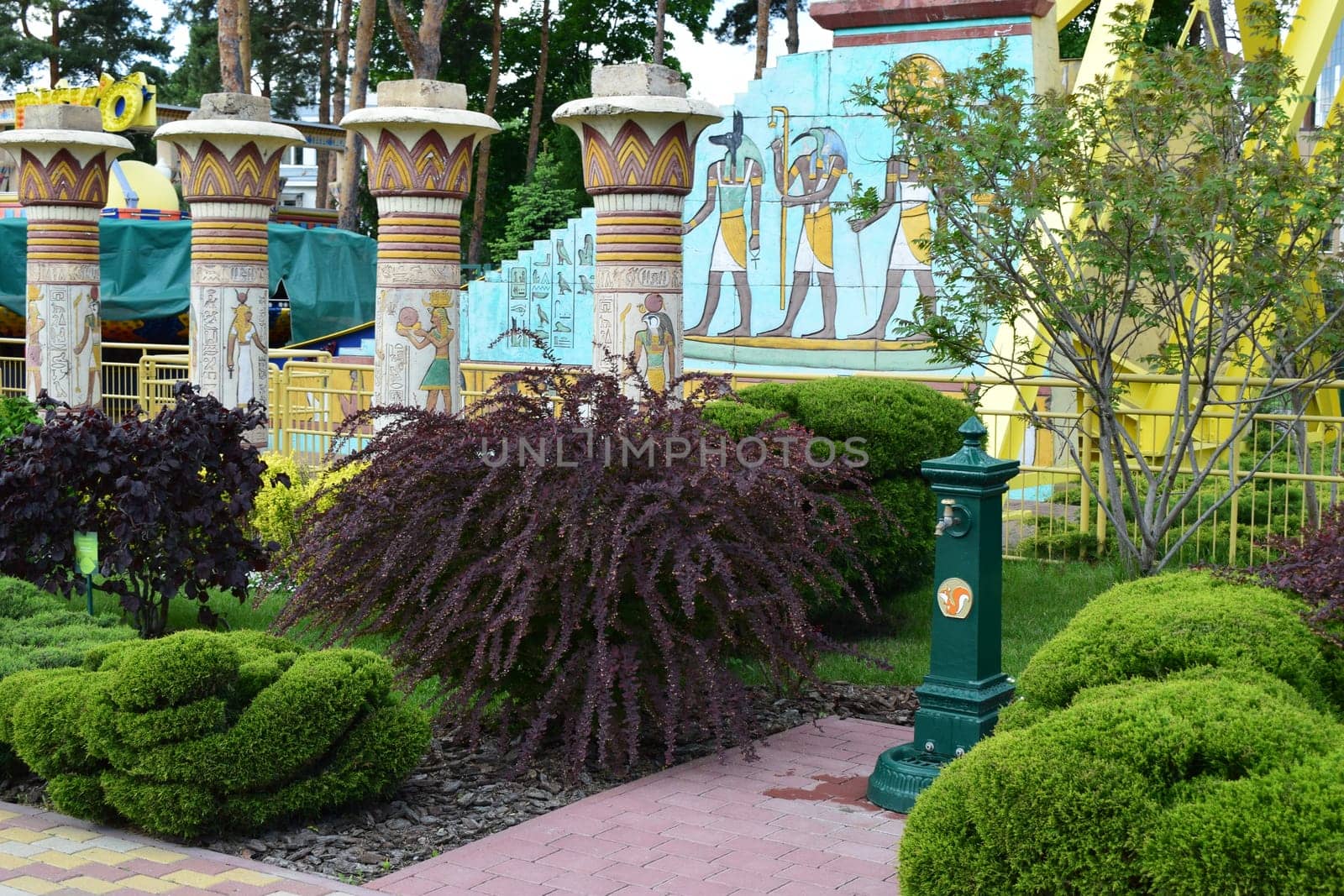 Kharkov, Ukraine, 14 May 2024. A themed park area with Egyptian-style columns and wall art depicting figures in traditional Egyptian attire. The area is landscaped with neatly trimmed bushes and trees, and there is a water fountain with a squirrel motif in front, set on a paved path. The surroundings include bright colors and playful designs, typical of an amusement park setting.
