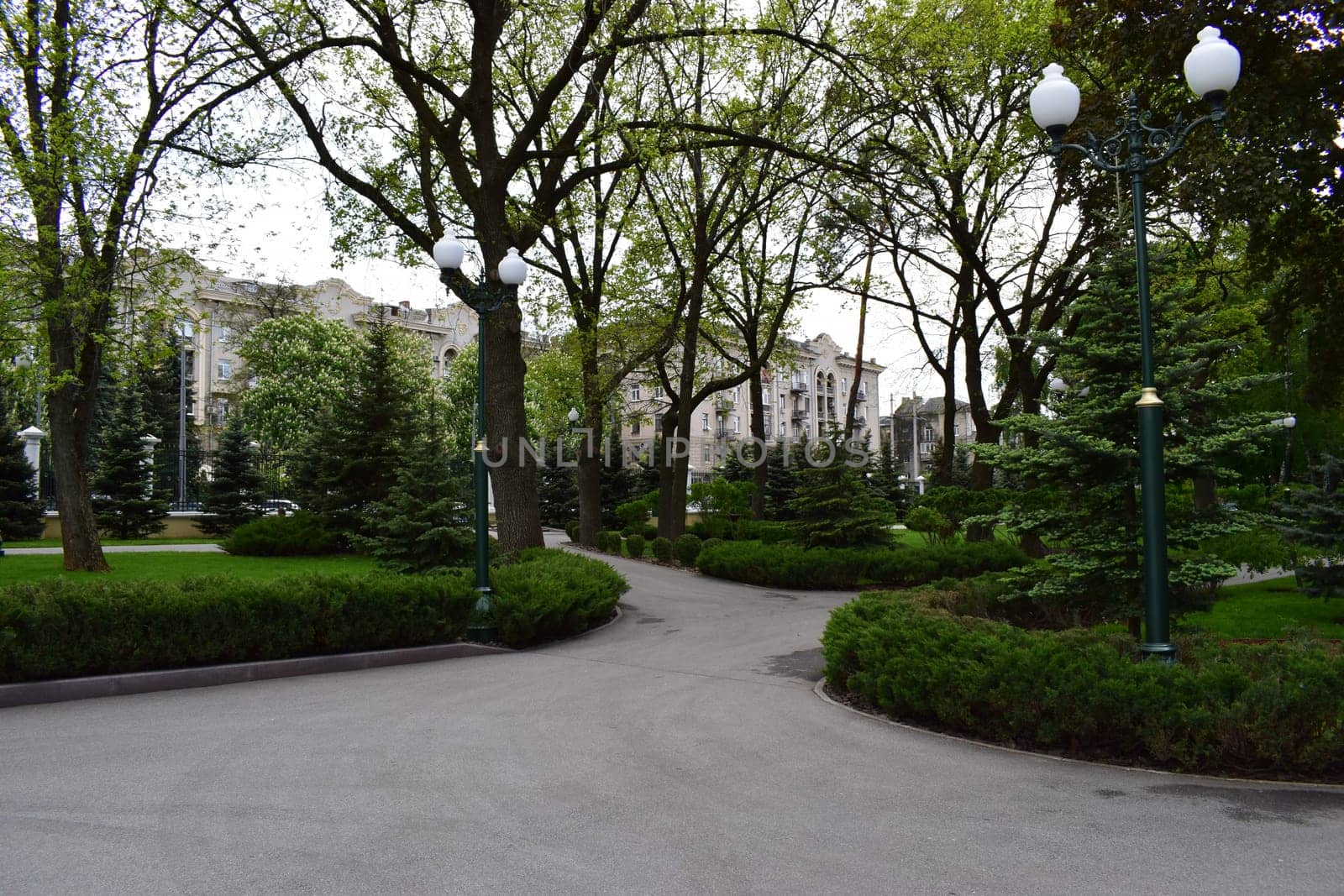Kharkov, Ukraine, 14 May 2024. An urban park with paved walkways lined with bushes and trees. There are several elegant lampposts and large residential buildings visible through the trees. The setting is green and well-kept, giving a sense of tranquility in the middle of the city.