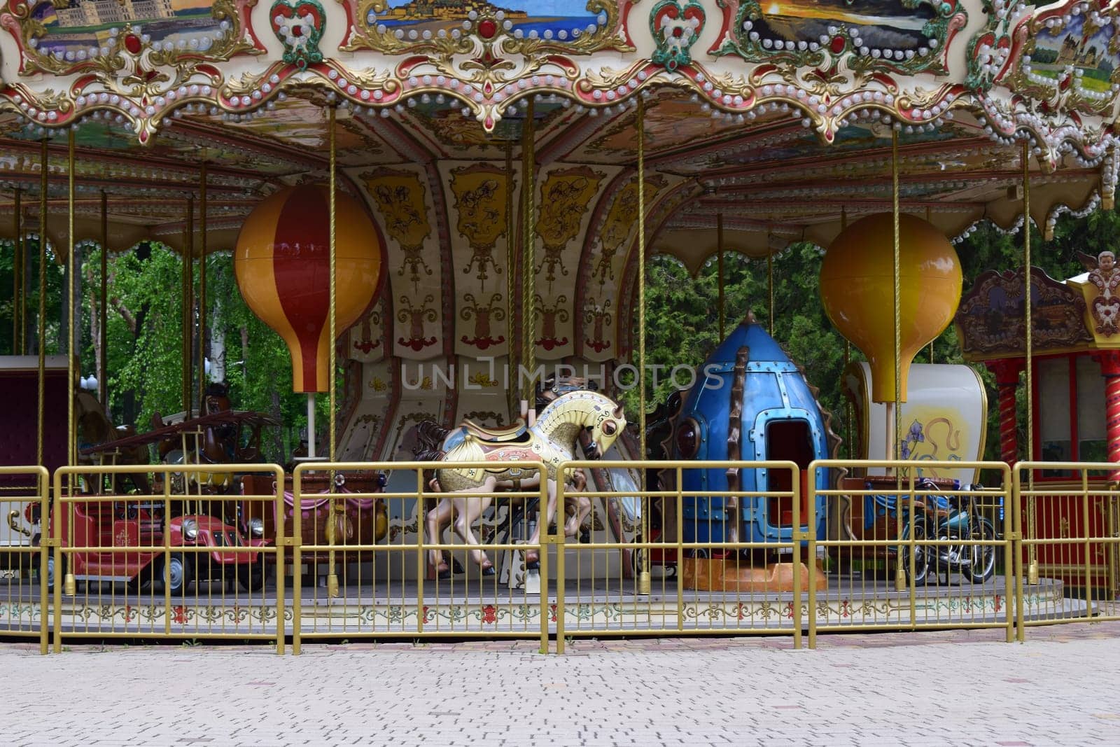 Kharkov, Ukraine, 14 May 2024. A vintage carousel with ornate decorations, featuring painted panels of scenic landscapes. The ride includes classic horses, a red car, and a blue rocket-shaped vehicle. Two large decorative spheres hang from the canopy, and the structure is enclosed by a yellow metal fence. The background shows green foliage, indicating an outdoor setting.