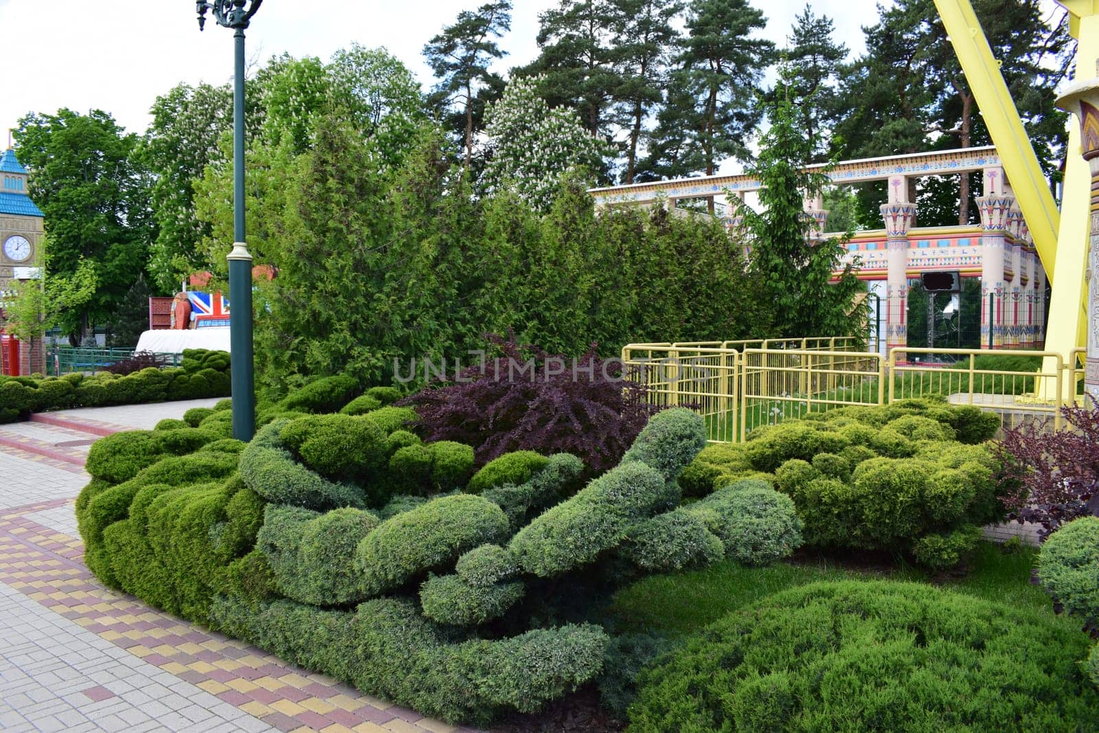 Kharkov, Ukraine, 14 May 2024. A garden scene with well-manicured greenery shaped into abstract topiary forms. In the background, there are ornate columns with Egyptian-style murals and decorations. The setting includes greenery, tiled pavement, and a colorful mural depicting Egyptian motifs.