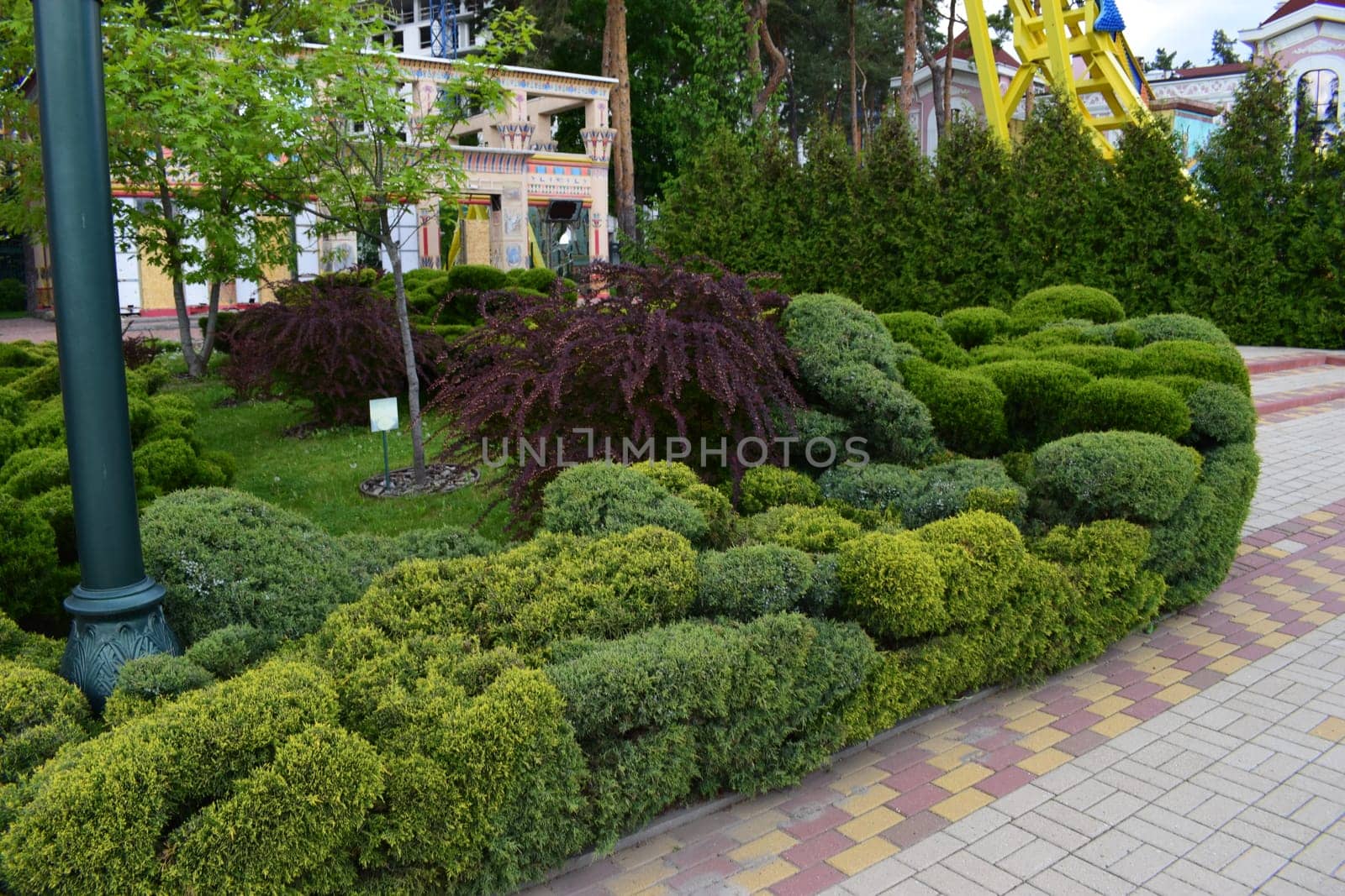 Kharkov, Ukraine, 14 May 2024. A well-kept garden with ornately trimmed shrubs and trees in the background. There is a brick path in shades of yellow and red and an architecturally designed structure in the background. A light pole is also visible on the left side of the image. In the background, an attractive yellow formation stands out among the vegetation.