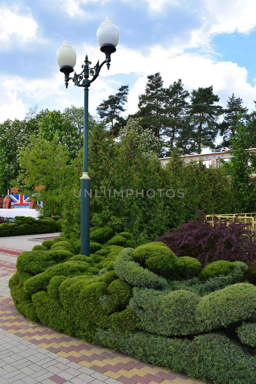 Kharkov, Ukraine, 14 May 2024. A landscaped garden scene featuring neatly trimmed greenery and decorative shrubs along a paved path. A vintage-style street lamp with dual globes stands prominently in the foreground. In the background, there's a clock tower resembling a classic European design, surrounded by lush trees under a partly cloudy sky.