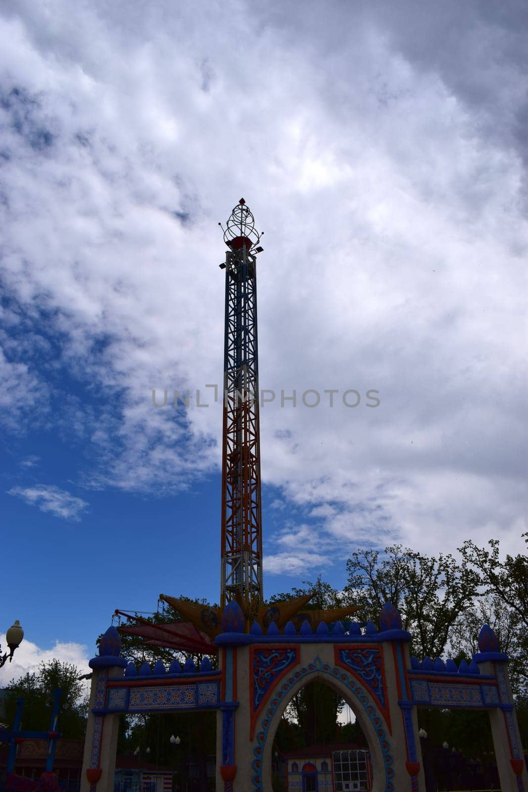Kharkov, Ukraine, 14 May 2024. A carousel tower at an amusement park. The structure is vertical and multicolored, with rich decoration at the base. The sky is partially obscured by clouds, with blue spots visible. The environment is green, with trees visible in the background.