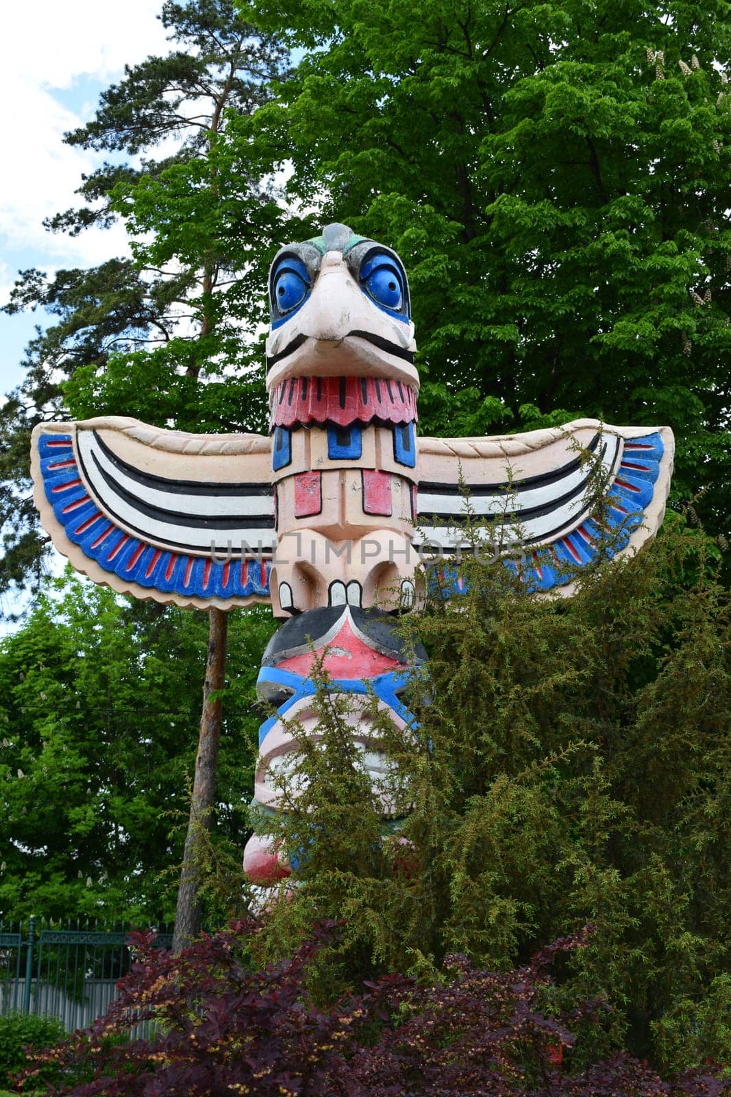 Kharkov, Ukraine, 14 May 2024. A large bird-shaped totem with bright colors, such as red, blue and white. It is located in a green environment, surrounded by trees and bushes under a partly cloudy sky.