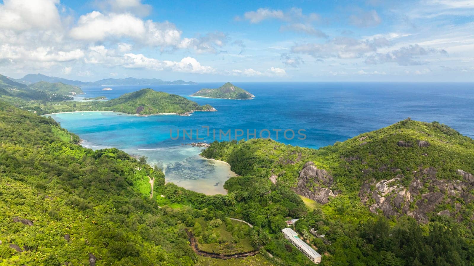 Coastal landscape with turquoise bays, lush green hills and distant islands under a cloudy sky. Seychelles, Mahe.