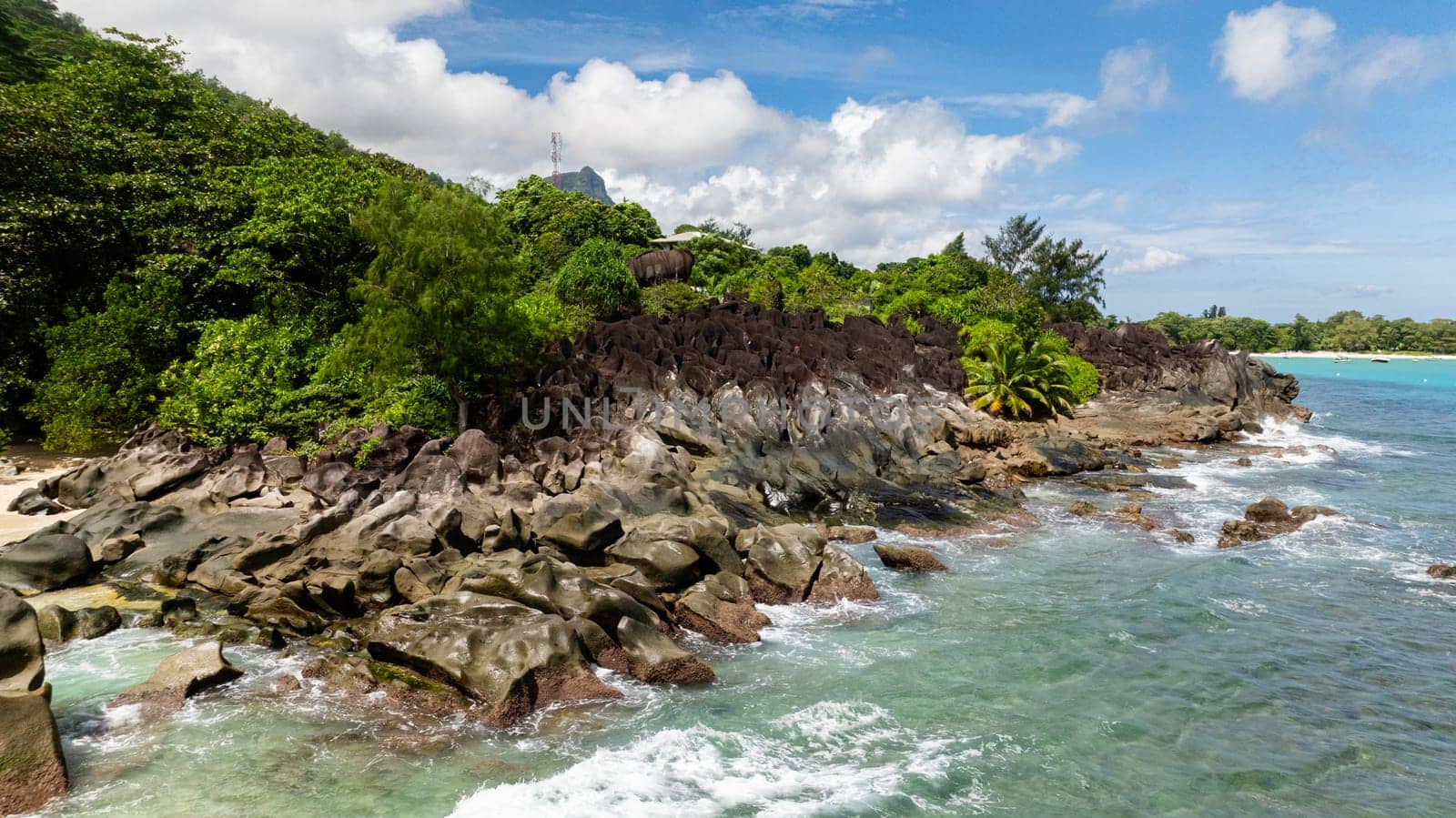 Rocky coastline with lush greenery and turquoise waters under a partly cloudy sky. Seychelles.