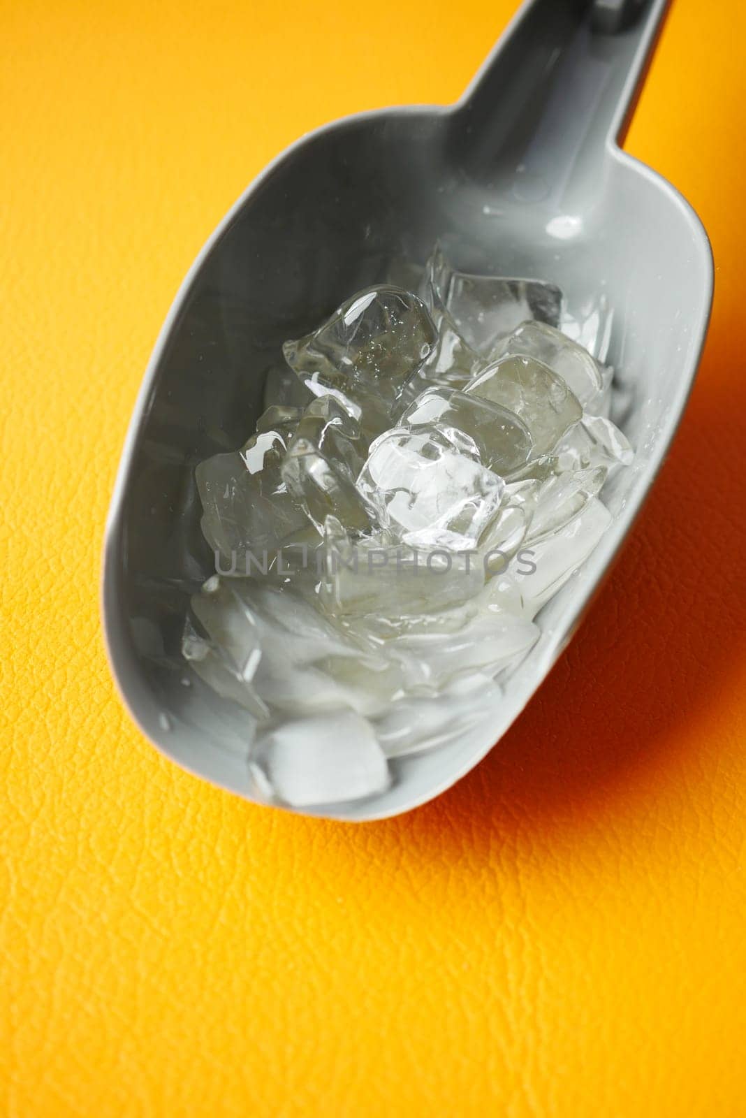 A scoop filled with clear ice cubes sits against a bright yellow backdrop.