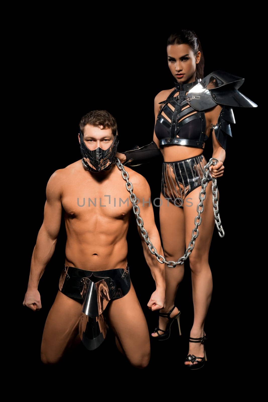 Kneeling muscular male in mask held on chain by dominant brunette female in black BDSM outfit on black background