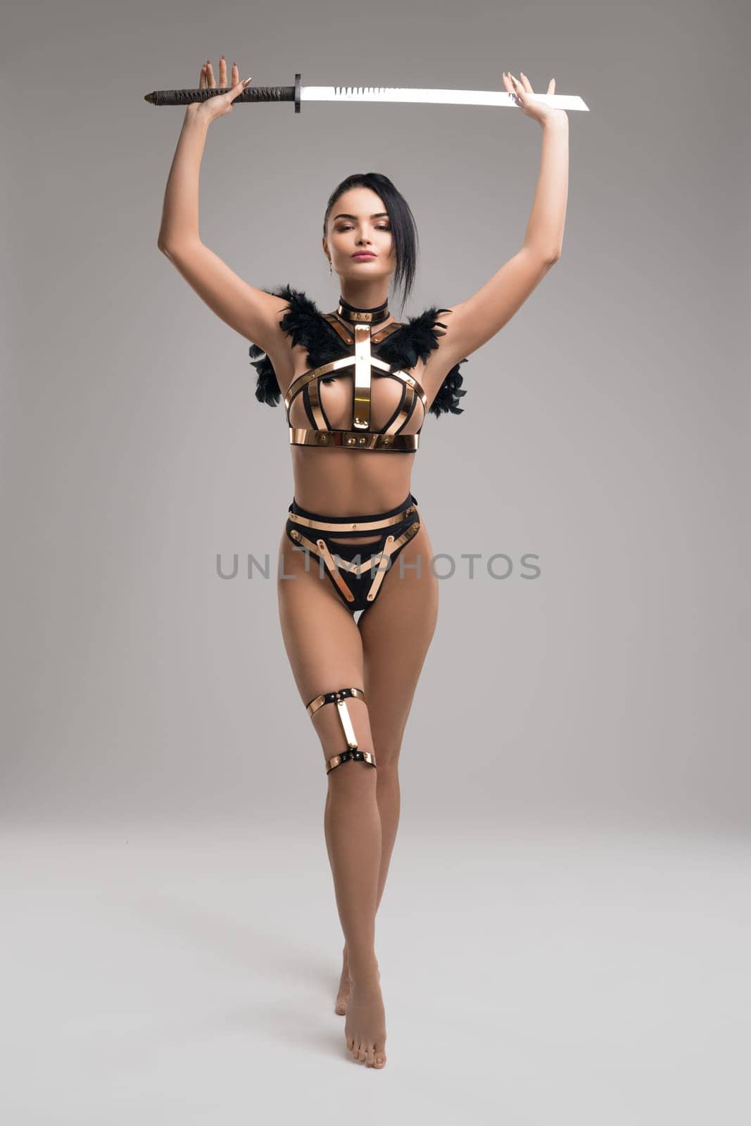 Full body sexy woman in armor shaped underwear stepping forward and carrying sharp sword in raised arms against gray background