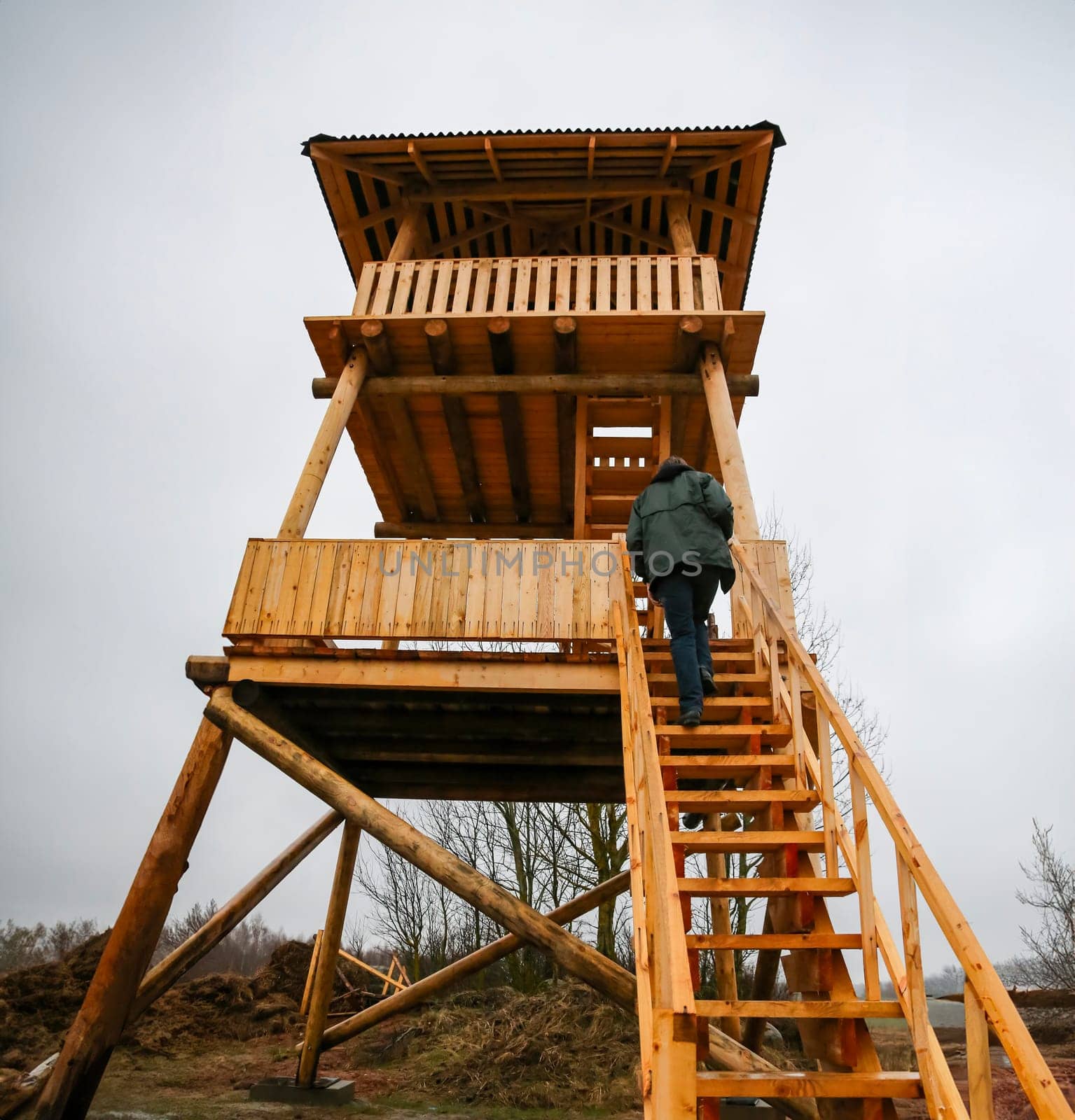A person climbs the wooden steps of a tall observation tower set in a natural landscape, under overcast skies, ready to enjoy the view.
