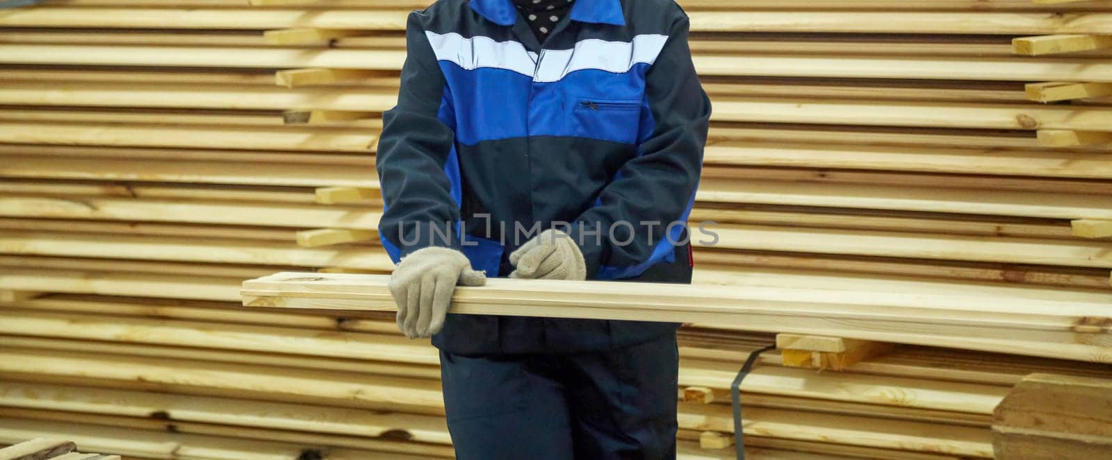A worker in blue and black attire uses a measuring tape on a wooden plank in a busy lumber yard filled with stacked timber.