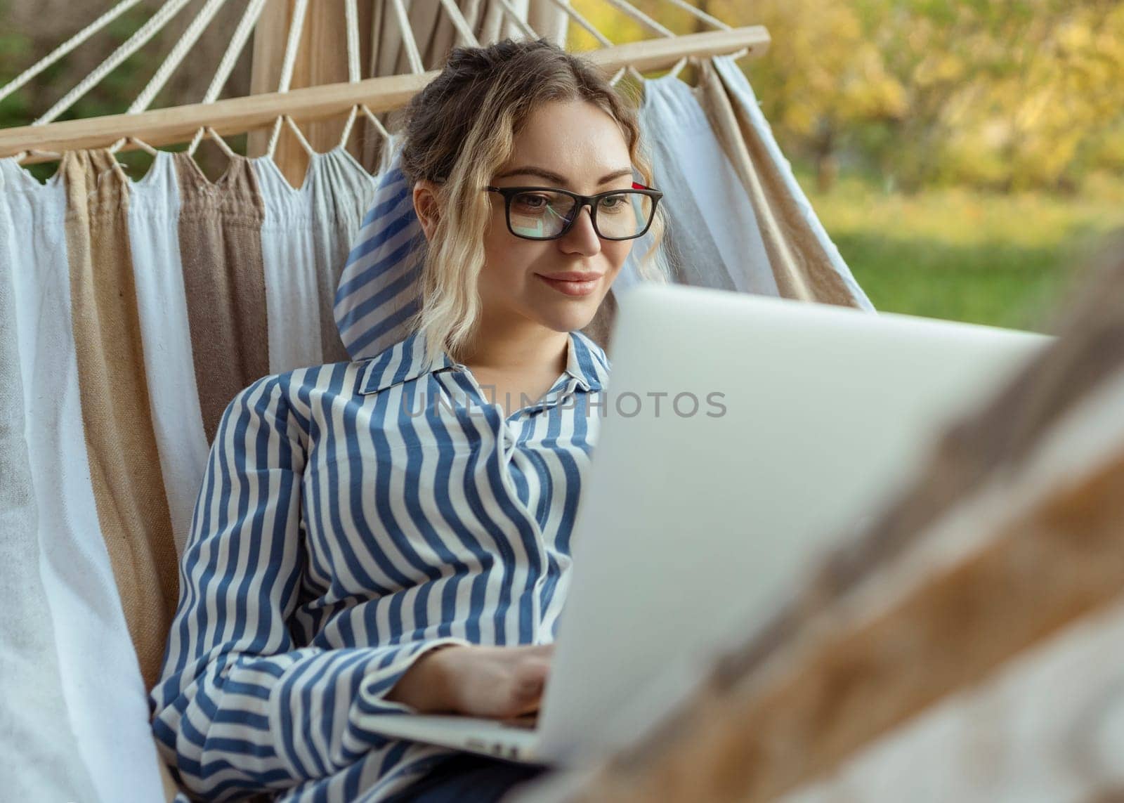 Young beautiful woman lies in a hammock with a laptop in glasses outside at home.