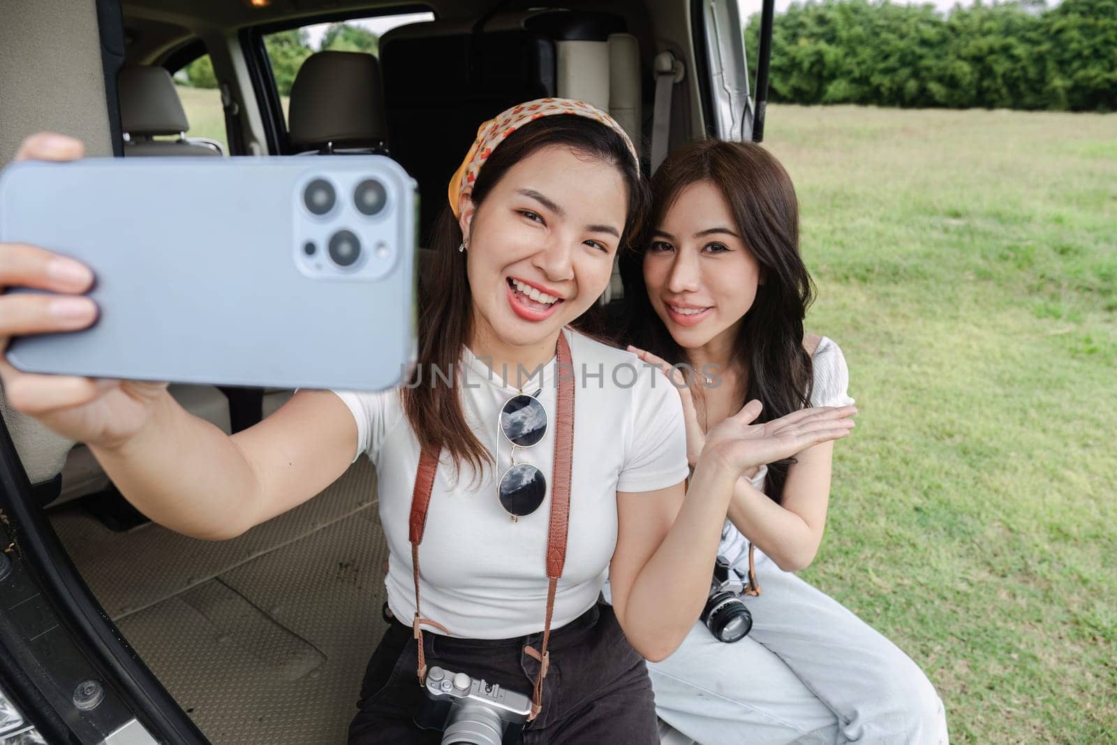 Two young women in white T-shirts and jeans sit back and relax, taking selfies together with their cell phones in the back of a car. by wichayada